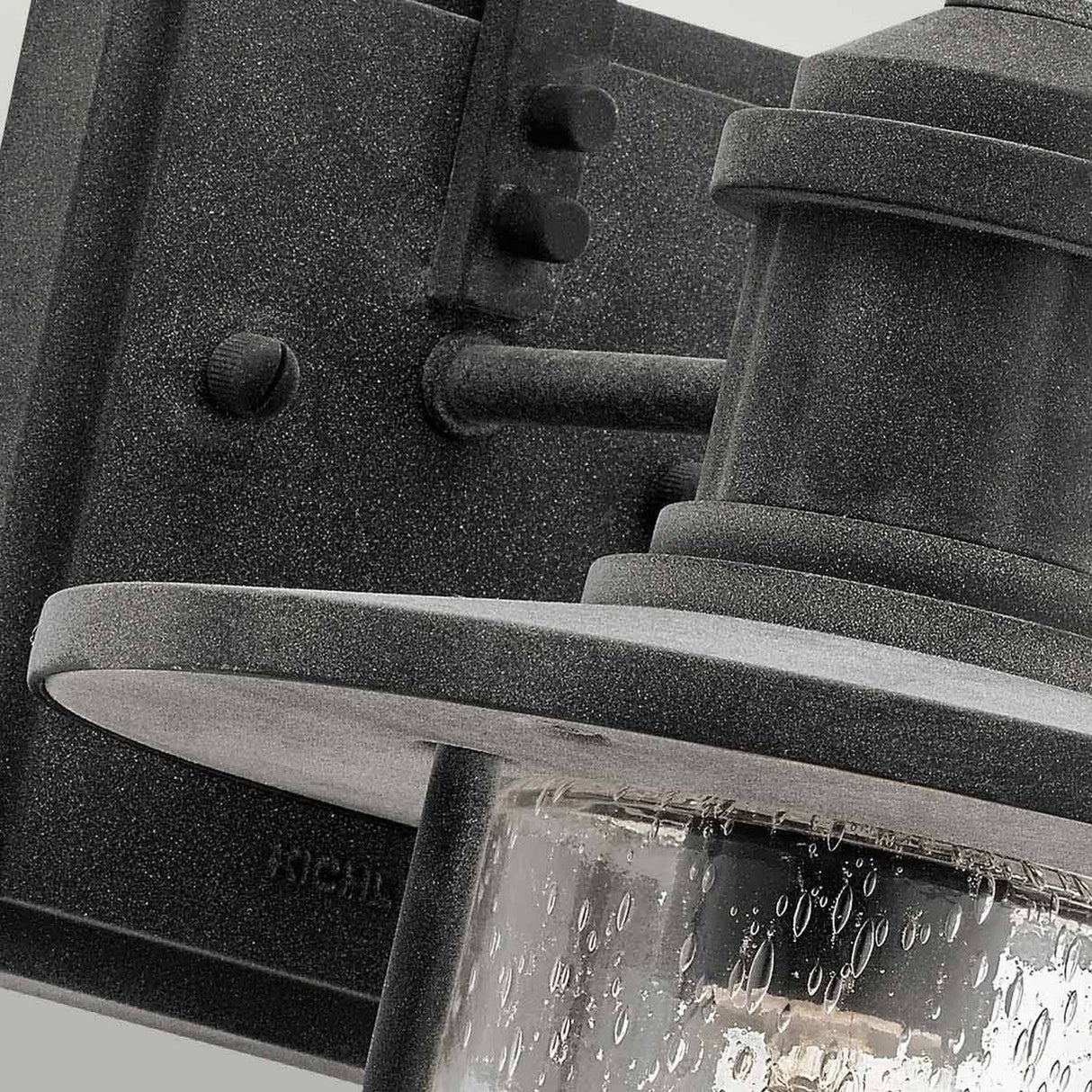 Close-up of the Riverwood Outdoor 1 Light Medium Wall Lantern, featuring a textured cylindrical clear glass shade. The fixture is affixed to a rectangular backplate crafted from weathered zinc, adding to its rustic elegance. The glass shade includes small water droplets for an added touch of charm.