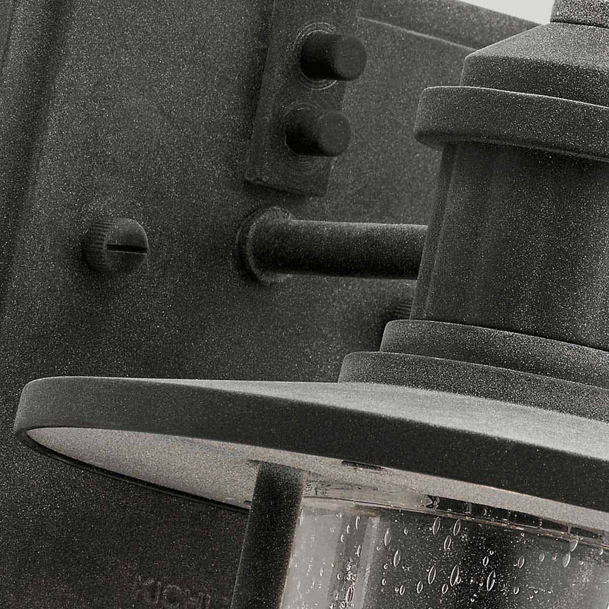 Close-up of the Riverwood Outdoor 1 Light Wall Lantern in weathered zinc, featuring a black finish and frosted glass cover that exudes a rustic seaside vibe. Raindrops cling to the glass, enhancing the nautical inspiration against the dark background.