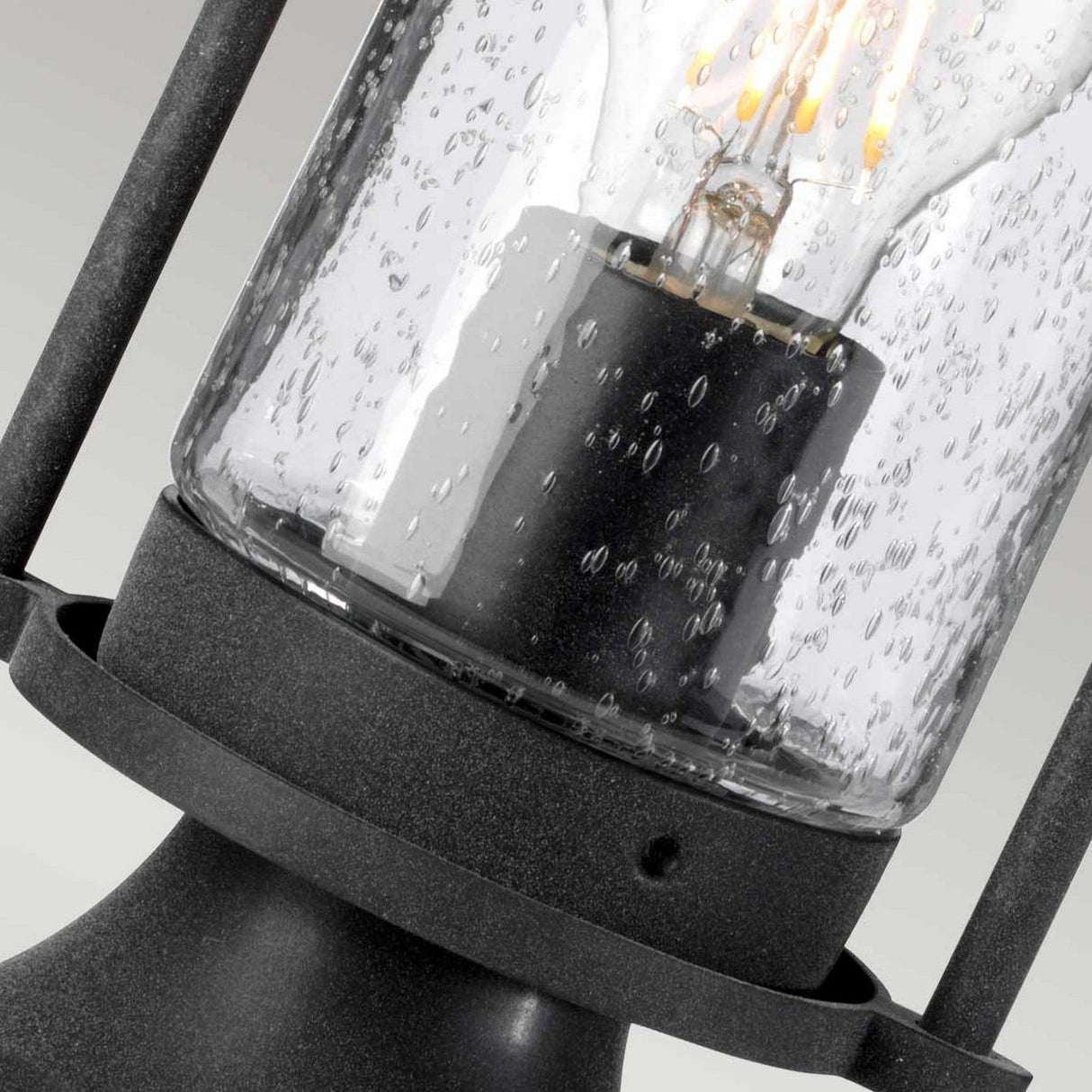 A close-up of the Riverwood Outdoor 1 Light Pedestal Lantern in weathered zinc reveals its black metal frame and clear glass cover with a visible light bulb inside. Its textured, bubbled surface creates a diffused lighting effect, while its nautical-inspired design enhances the charm against the neutral backdrop.