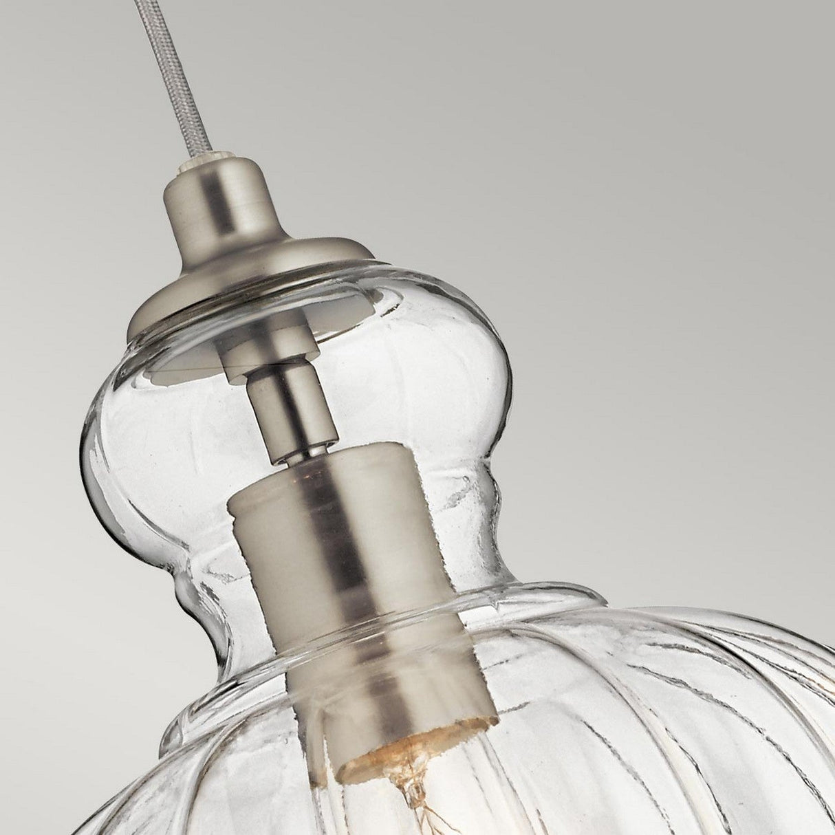 A close-up of the Riviera-1 1 Light Pendant highlights its clear, textured dome-shaped shade with vintage-inspired elegance. The fixture includes a brushed nickel metal fitting and a visible bulb inside, connected by a cord against a neutral gray background.