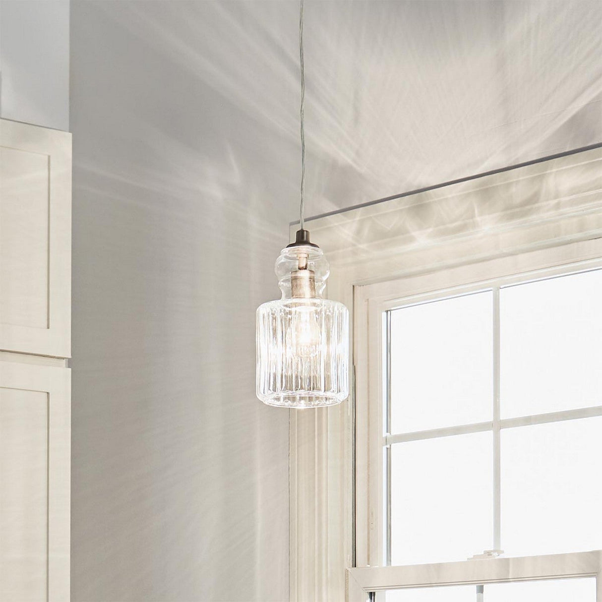 The Riviera-2 1 Light Pendant in brushed nickel hangs from the ceiling beside a window, featuring a clear glass shade. Sunlight filters through the glass, casting patterns on the wall. Ideal for kitchen island lighting, this setup complements the room's minimalist design and light-colored walls.