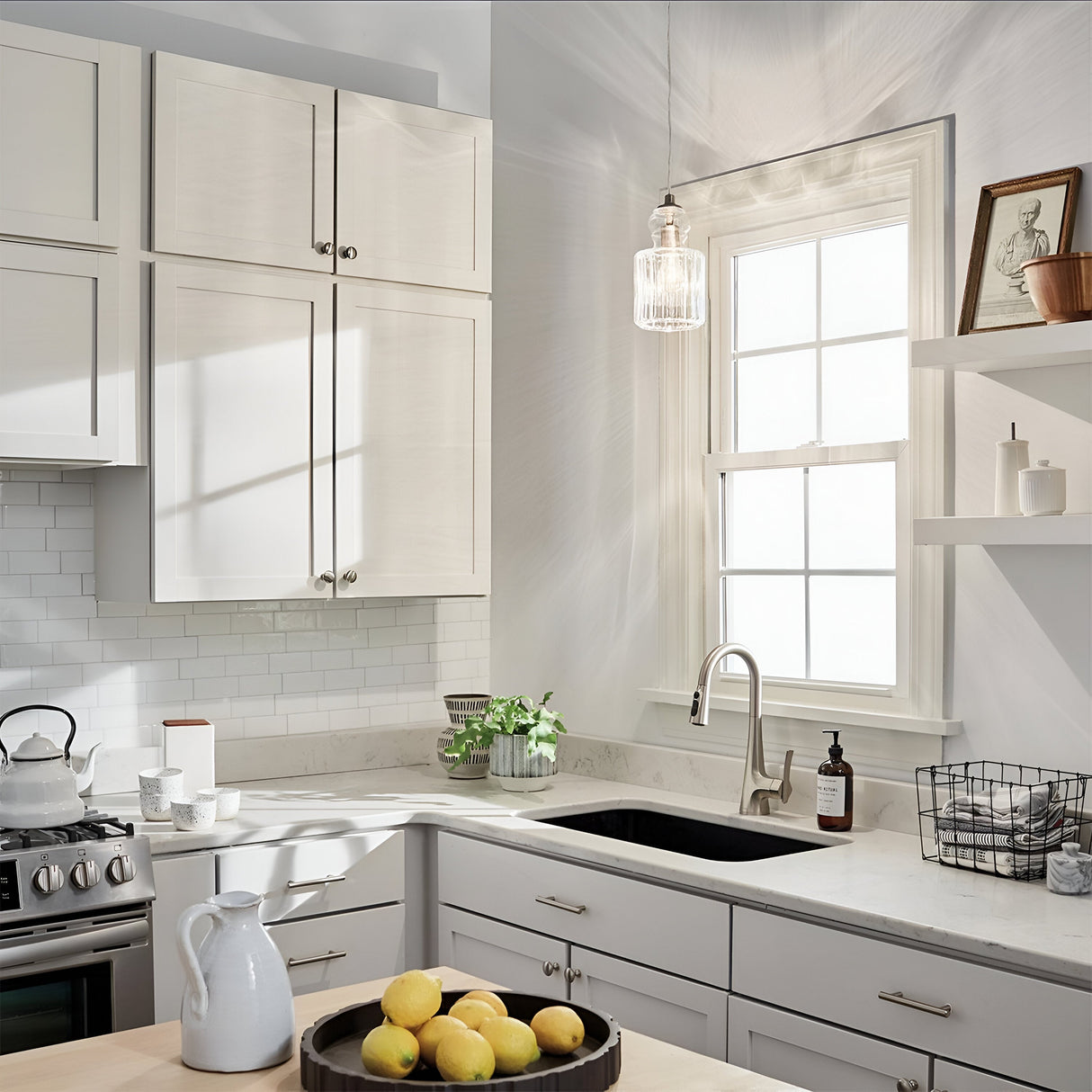 The sunlit kitchen features white cabinets, a silver faucet, and a black sink. The Riviera-2 1 Light Pendant in brushed nickel hangs gracefully above. A bowl of lemons on the counter enhances the ambiance, while a stove, kettle, and green plant add decorative touches. Framed art and bottles rest on shelves near a window with white trim.