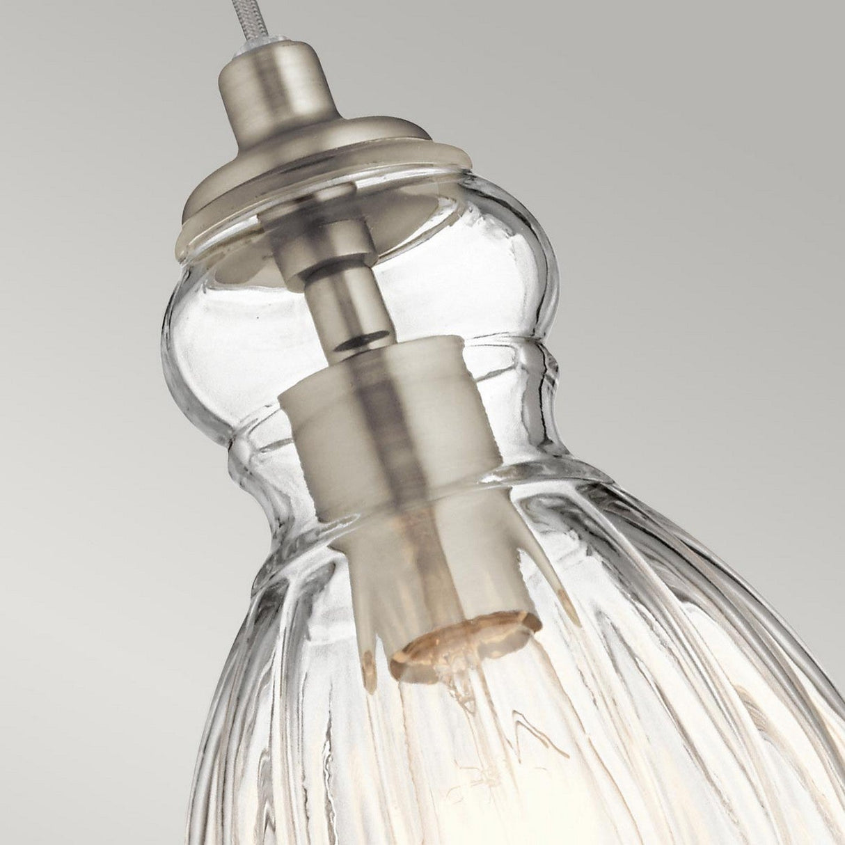 Close-up of the Riviera-3 1 Light Pendant, featuring a vintage-inspired design with a clear ribbed glass shade and brushed nickel top. The partially visible light bulb inside is set against a neutral gray background.