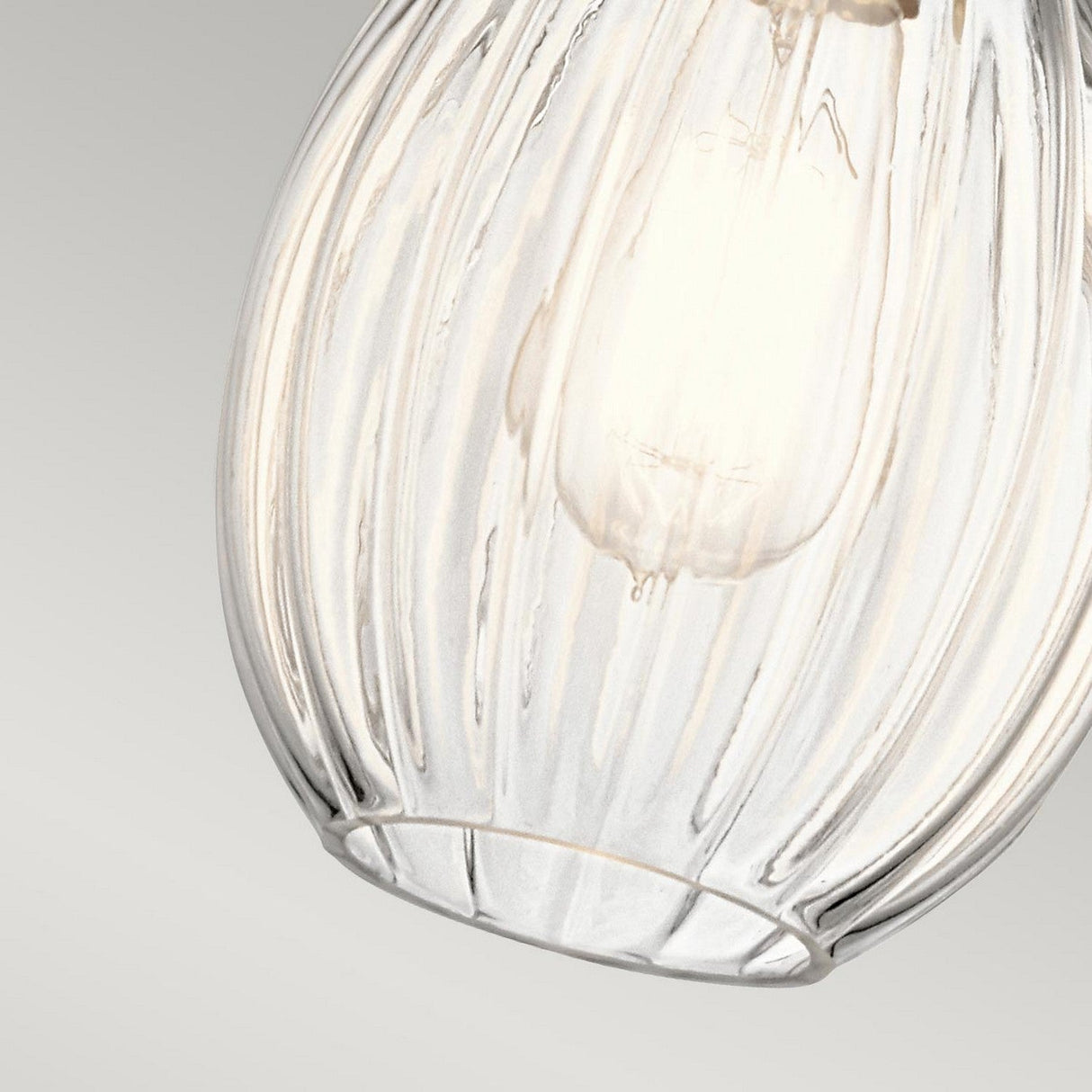 A detailed view of the Riviera-3 1 Light Pendant – Brushed Nickel showcases its vintage-inspired design with a clear, ribbed glass finish and an illuminated bulb inside. The background features a gradient of light gray tones, highlighting the brushed nickel accents and elegant simplicity of the fixture.