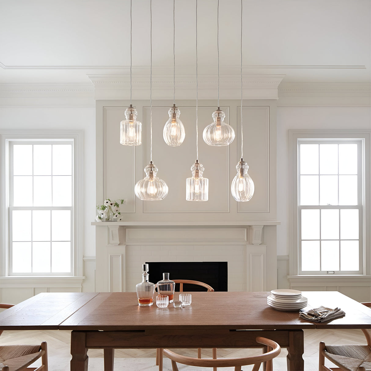 A dining room with a wooden table set for a meal, featuring plates and glasses. Hanging above the table are six Riviera-3 1 Light Pendants in brushed nickel, each with a vintage-inspired design and glass shade. In the background, there is a white fireplace and two large windows that contribute to the room's bright, airy feel.
