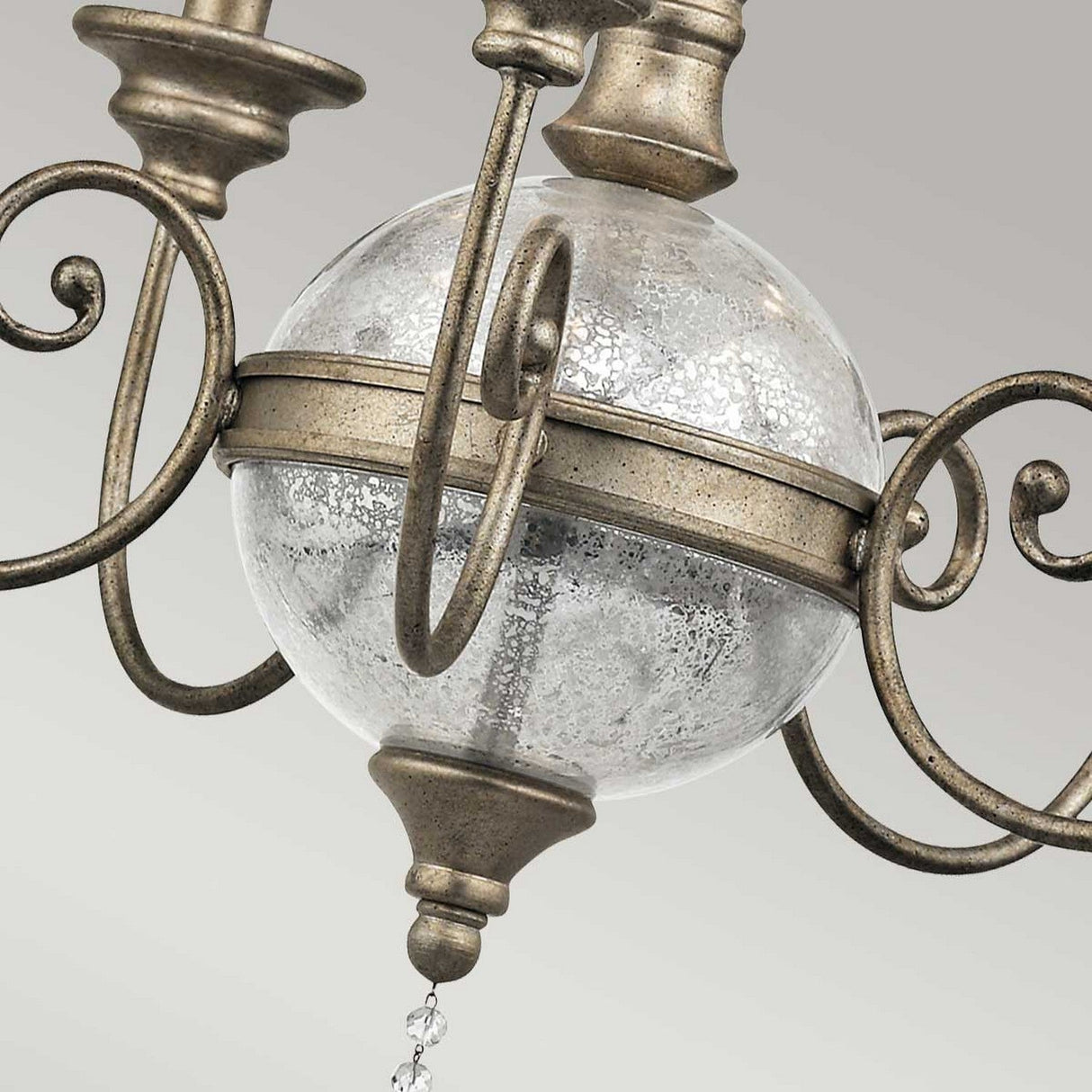 Close-up of the intricate details on the Rosalie 5 Light Chandelier in a classic design. It features a textured antique mercury glass globe at the center and intricate metal scrollwork in a sterling gold finish. The background is a soft gray, enhancing its elegant allure.