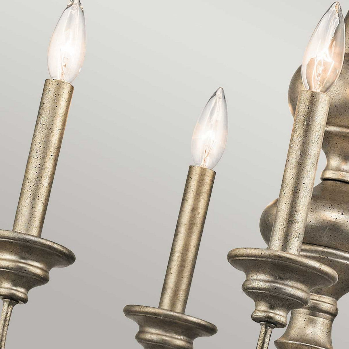 Close-up of the Rosalie 5 Light Chandelier in Sterling Gold featuring five candle-shaped bulbs in metallic holders. The bulbs emit a warm glow against a light gray background, highlighting the elegant Williamsburg design and structure of this exquisite fixture.