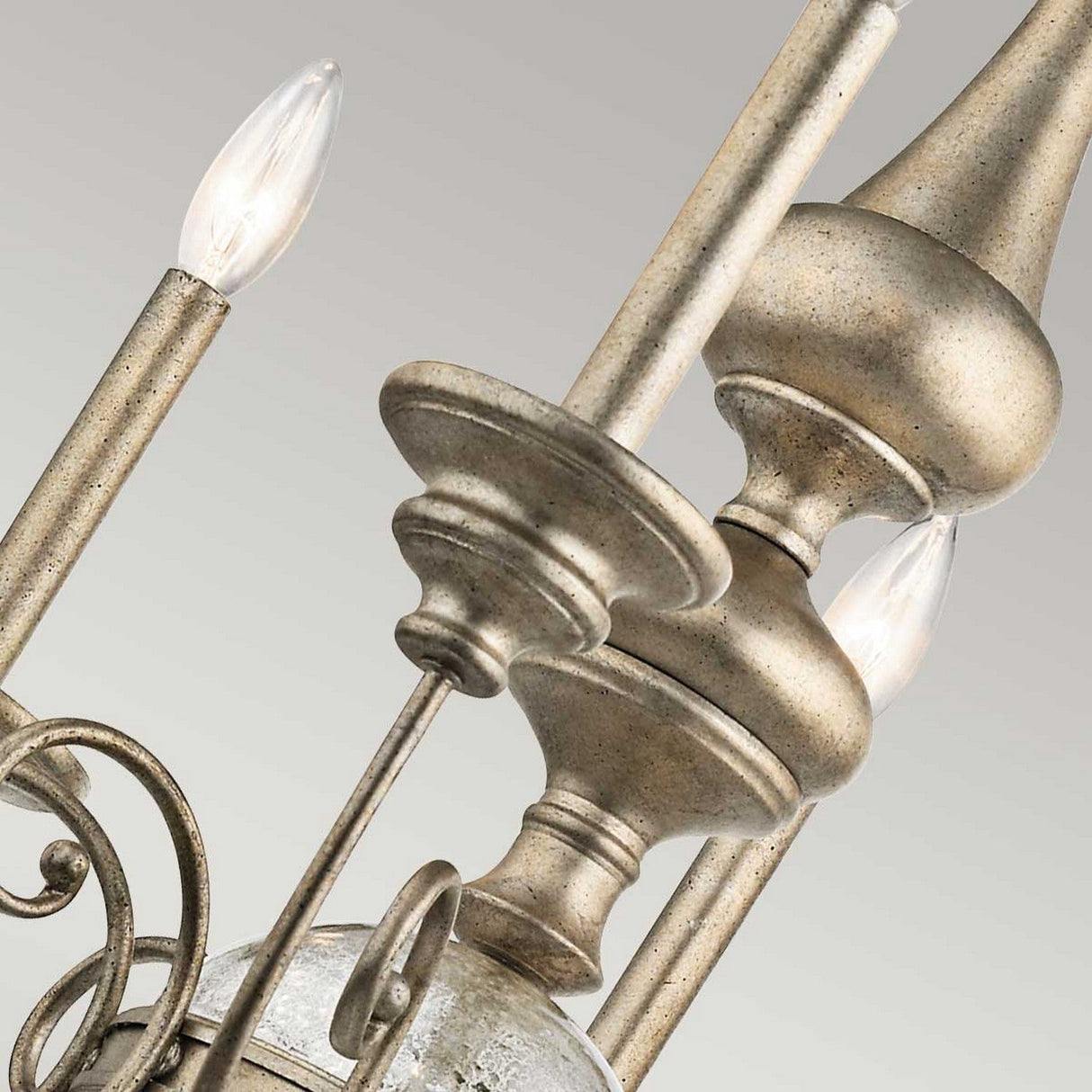 A close-up of the Rosalie 8 Light Chandelier in Sterling Gold displays candle-shaped light bulbs on slender stands surrounded by intricate metalwork, echoing the charm of Antique Mercury Glass. The elegant design is accentuated against a plain, light-colored background.