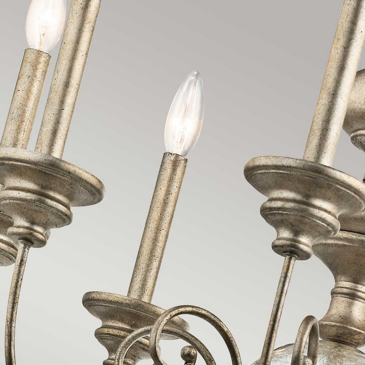 Close-up of the Rosalie 8 Light Chandelier in Sterling Gold, featuring candle-shaped bulbs and intricate metal detailing against a light gray background. The fixture, echoing Williamsburg design, showcases a rustic, weathered finish.