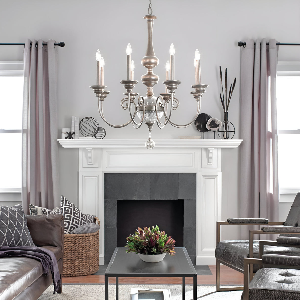 A cozy living room showcases a Rosalie 8 Light Chandelier in Sterling Gold, complemented by a white fireplace with slate accents and modern decor. Two windows with sheer curtains frame the fireplace. A coffee table features a plant surrounded by patterned cushions and chairs, adding Williamsburg design charm.