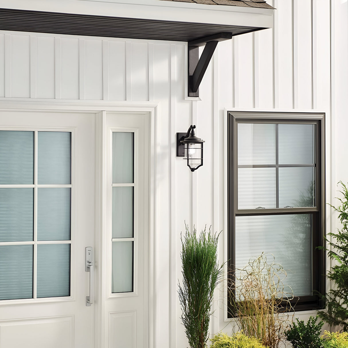 A minimalist house exterior showcasing a white door with vertical seeded glass panels is complemented by a window with a black frame and grid design. Above the small garden filled with various plants, enhancing the nautical aesthetic, is mounted the Royal Marine Outdoor 1 Light Small Wall Lantern in black.
