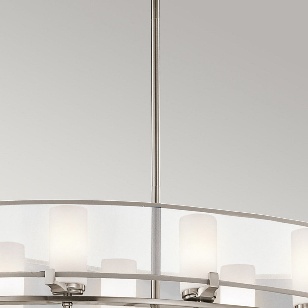 Close-up of the Saldana 8 Light Oval Chandelier - Silver, showcasing its celestial-inspired design with a brushed metal finish. The lighting fixture features frosted glass cylindrical shades arranged in a circular pattern, casting a soft glow against the plain, light-colored wall.