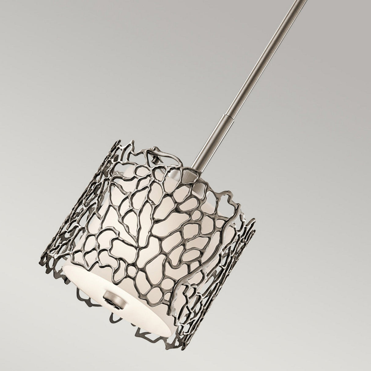 The Silver Coral 1 Light Mini Pendant - Silver offers a contemporary vibe with its modern design, showcasing a cylindrical white lampshade enclosed within an openwork metal frame. The abstract web pattern adds a nature-inspired elegance, beautifully suspended from a slender metal rod.