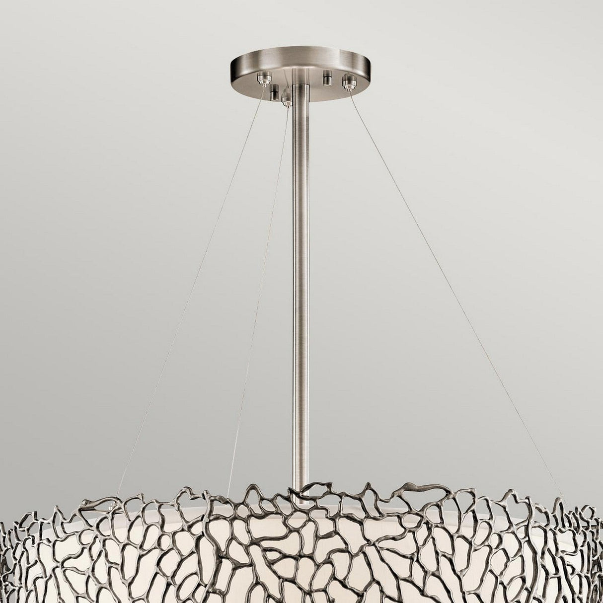 Introducing the Silver Coral 4 Light Drum Pendant - Silver, a modern ceiling light fixture that exudes elegance with its metallic abstract design inspired by nature. This chandelier showcases a metal frame adorned with a wavy, coral-inspired pattern encircling a circular white shade, all supported by a central rod and wires against a plain, light-colored wall.