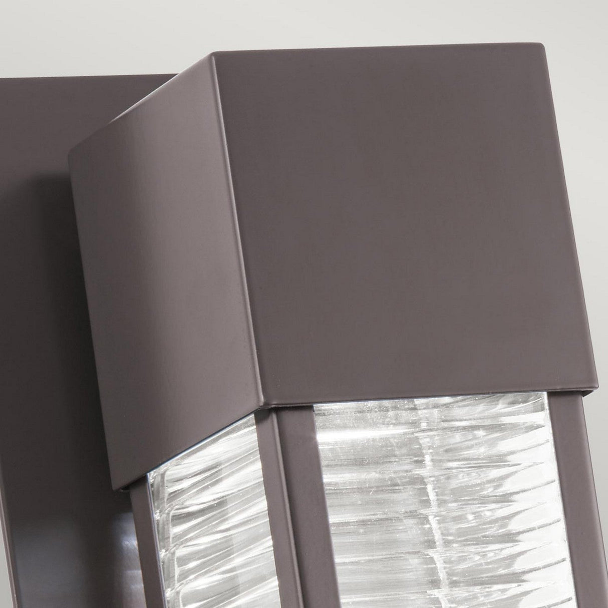 Close-up of the Sorel Outdoor 1 Light Wall Lantern - Bronze, featuring a sleek, square design. The lantern showcases an Architectural Bronze finish and clear glass panels for a contemporary appearance. Its surface emits a soft glow, accentuating the textured details.