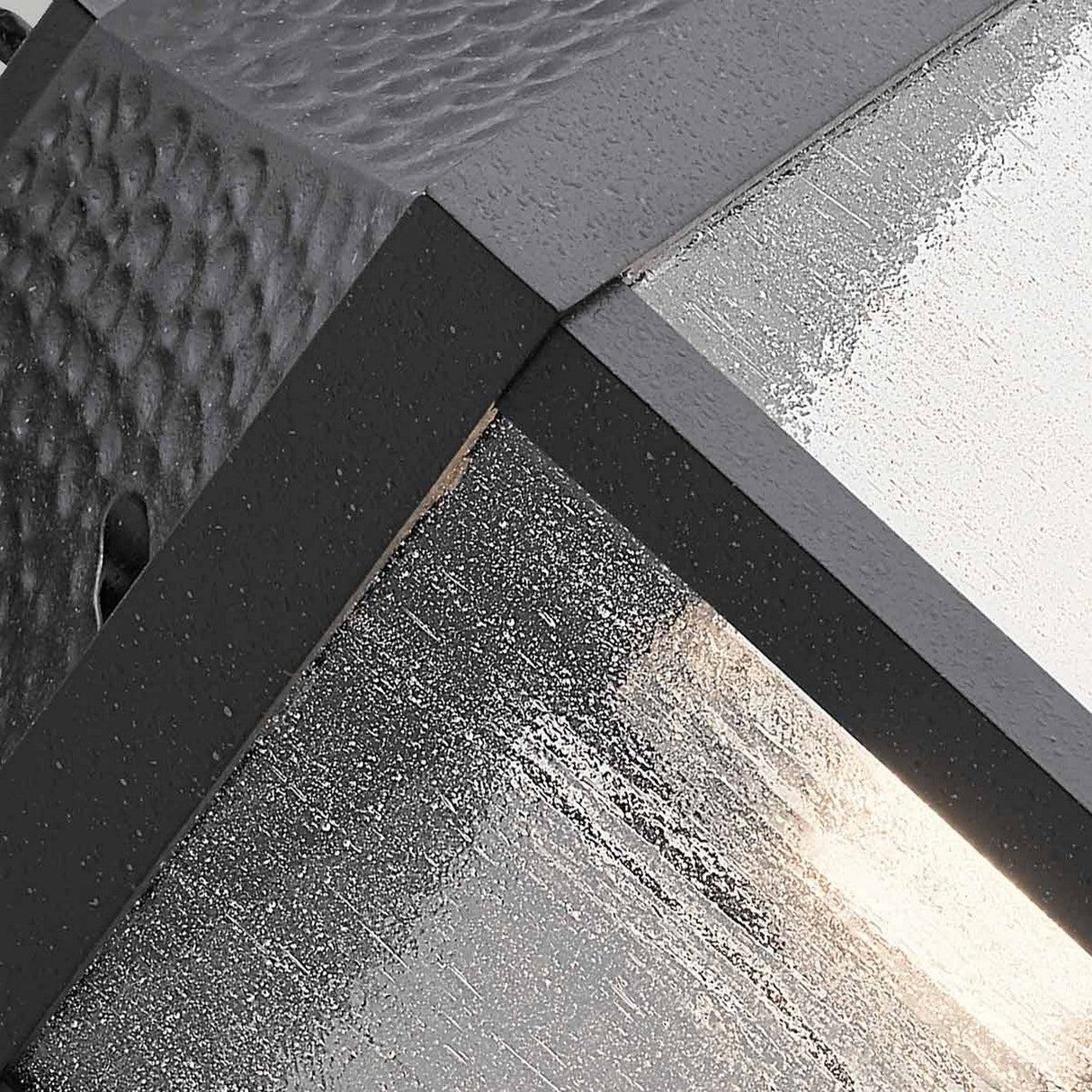 Close-up view of the Springfield Outdoor 1 Light Medium Wall Lantern in black, showcasing its modern design with textured black metal and frosted glass. The distressed finish highlights the intersection of panels, featuring a combination of smooth and hammered metal textures that evoke an outdoor wall lantern aesthetic.
