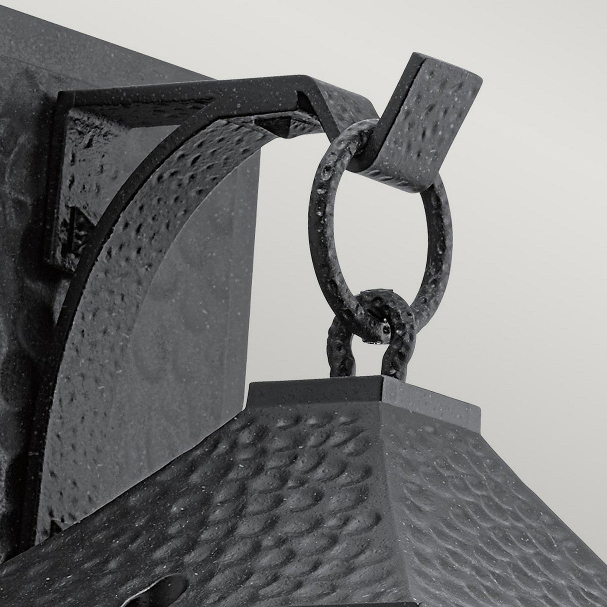 Close-up of the Springfield Outdoor 1 Light Medium Wall Lantern in a distressed black finish with a hammered texture. This lantern is suspended from a decorative bracket featuring a ring and hook design, complemented by seeded glass that enhances its charm. The plain light grey background accentuates the lantern’s intricate details and craftsmanship.