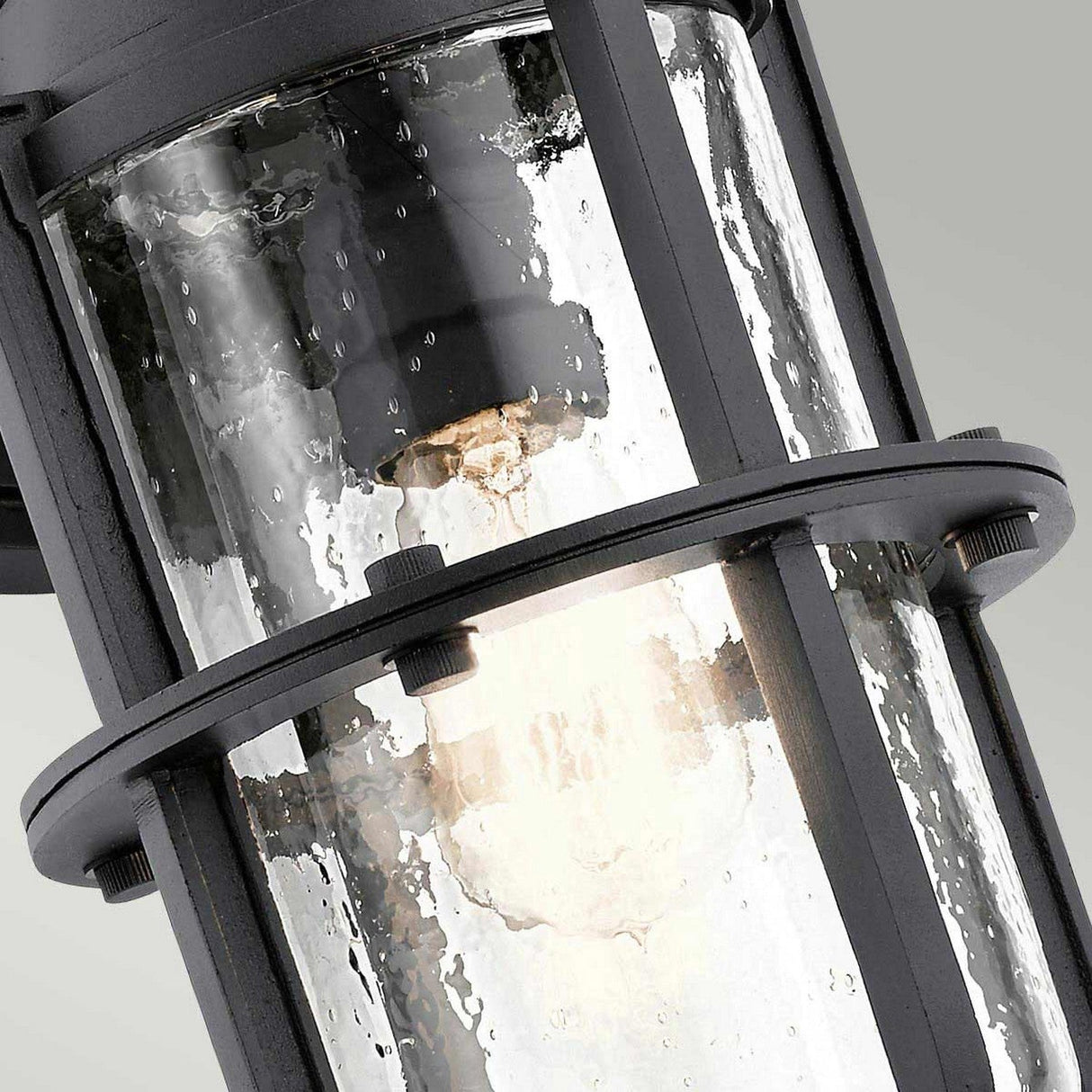 Close-up of the Suri Outdoor 1 Light Medium Wall Lantern in black, showcasing its coastal-inspired cylindrical design. It features transparent, textured glass encasing a glowing light bulb inside. The fixture's dark metal frame, supported by horizontal and vertical bars, enhances durability and charm while providing UV protection.