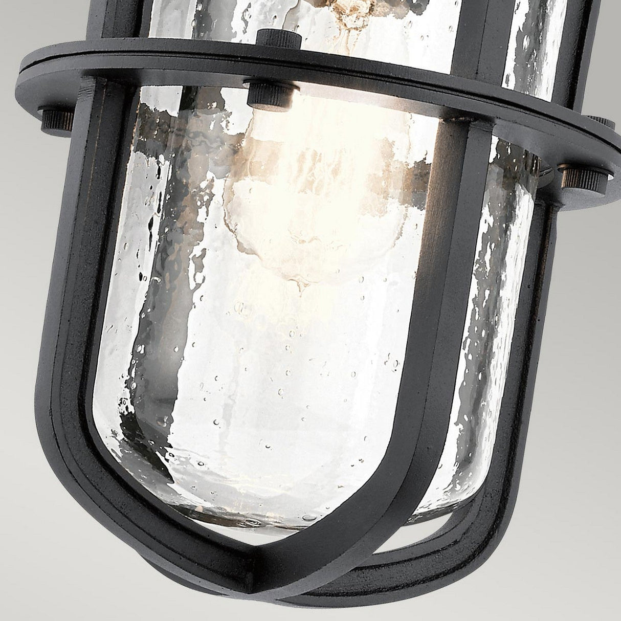 Close-up of the Suri Outdoor 1 Light Medium Wall Lantern in black, featuring an industrial design with a clear glass shade and metal cage reminiscent of coastal-inspired lighting. The textured glass bulb is illuminated, casting a warm glow against a neutral background.
