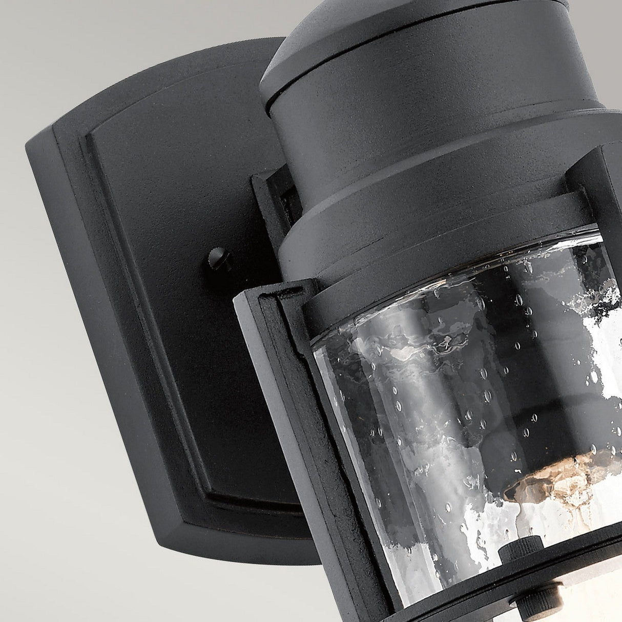 Close-up of the Suri Outdoor 1 Light Medium Wall Lantern in black, showcasing a glass cylinder with bubble details. The fixture is mounted on a rectangular base, providing coastal-inspired lighting and featuring a sleek metal finish.