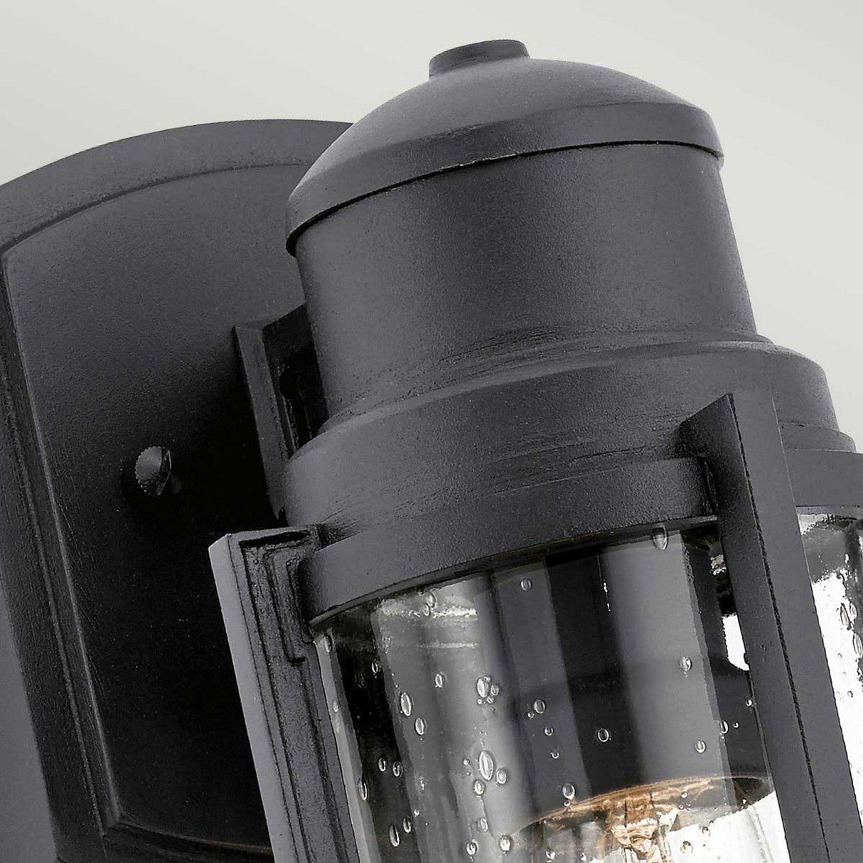 The Suri Outdoor 1 Light Small Wall Lantern in black features a sleek and modern design with a clear cylindrical glass cover. Mounted against a light gray background, the minimalist metal structure shines, with water droplets visible on the glass surface to accentuate its elegant coastal aesthetic.