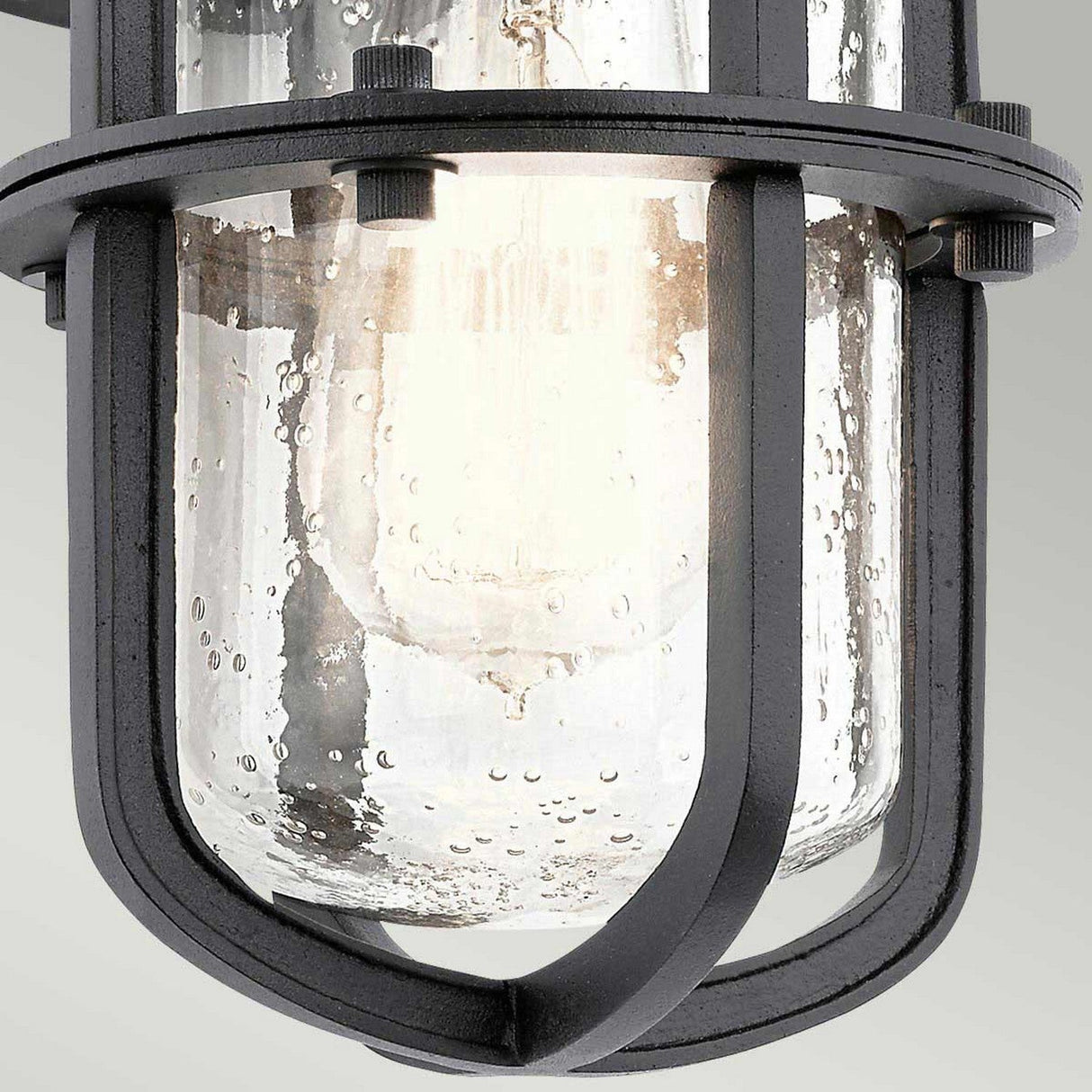 Close-up of the Suri Outdoor 1 Light Small Wall Lantern in black, exuding a vintage industrial style. It showcases a cylindrical glass cover encased in a metal cage, with its speckled texture gently revealing the bulb inside and casting a warm glow reminiscent of coastal design lanterns.