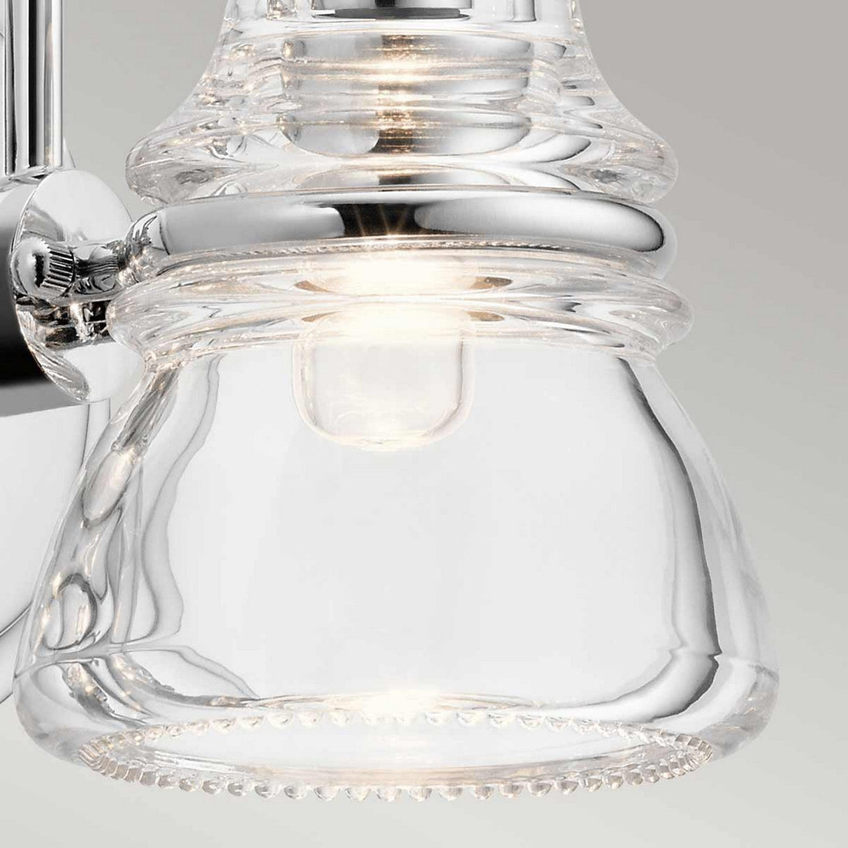 The Talland 1 Light Wall Light - Polished Chrome features prominently in the close-up, highlighting its polished chrome details that offer a vintage aesthetic. The light is illuminated, casting a gentle glow through the clear glass against a simple gray background.