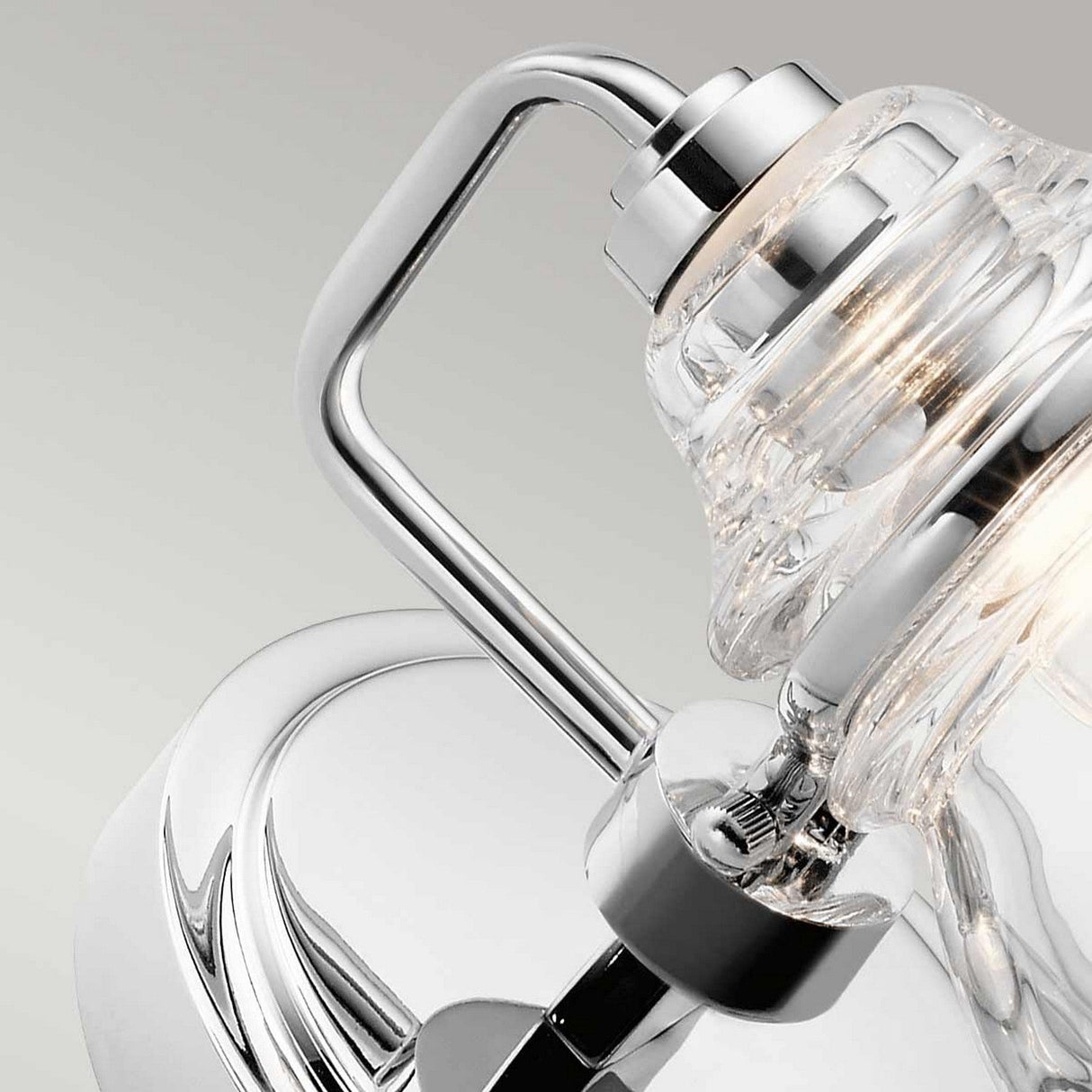 Close-up of the Talland 1 Light Wall Light - Polished Chrome, featuring a clear glass shade that showcases the visible light bulb, casting a soft glow. The wall light's sleek polished chrome finish enhances its curved lines, exuding a vintage aesthetic against a plain background.