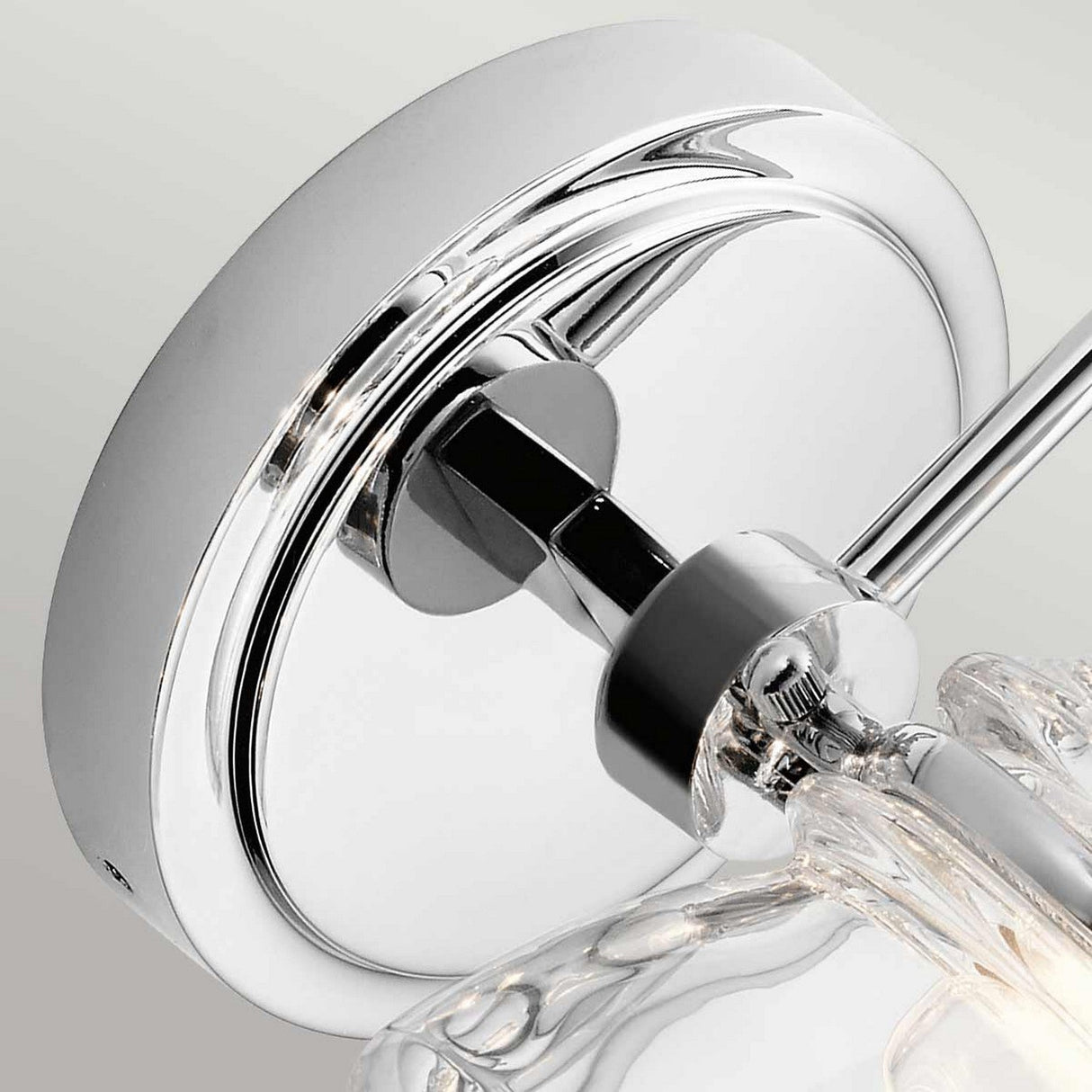 A close-up of the Talland 1 Light Wall Light - Polished Chrome showcases its circular wall mount and clear glass component, highlighting its sleek design that beautifully reflects light. The fixture combines modern style with a subtle vintage touch against a neutral background.