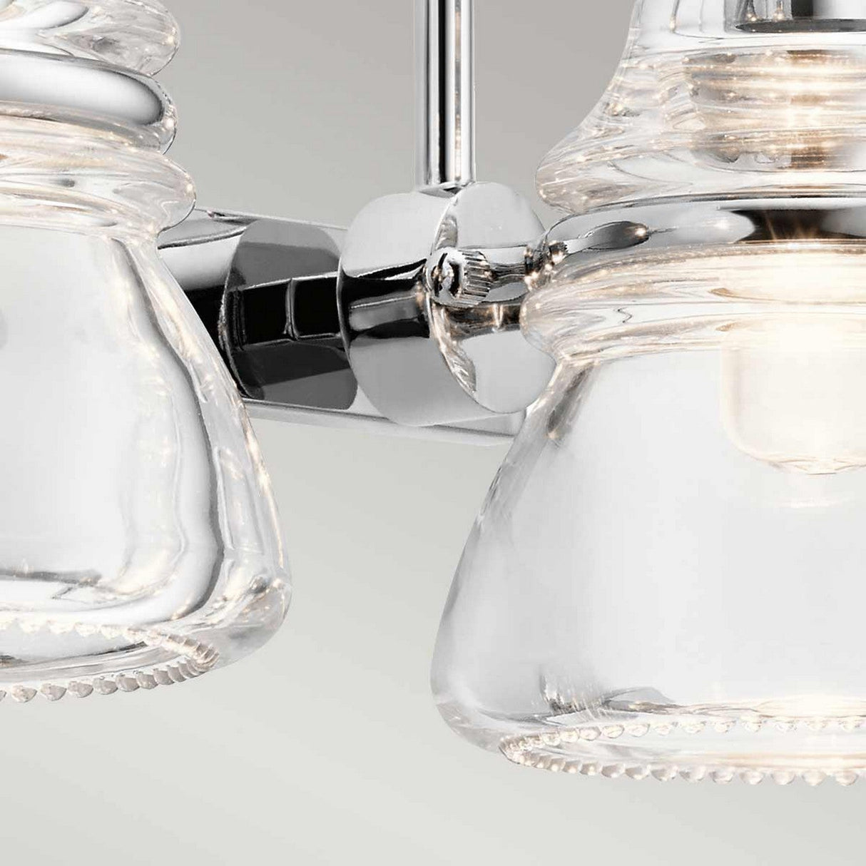 A close-up of the Talland 2 Light Wall Light in polished chrome showcases its modern design with two clear glass shades, vintage-inspired glasswork, and intricate details. It features a polished finish against a neutral background.