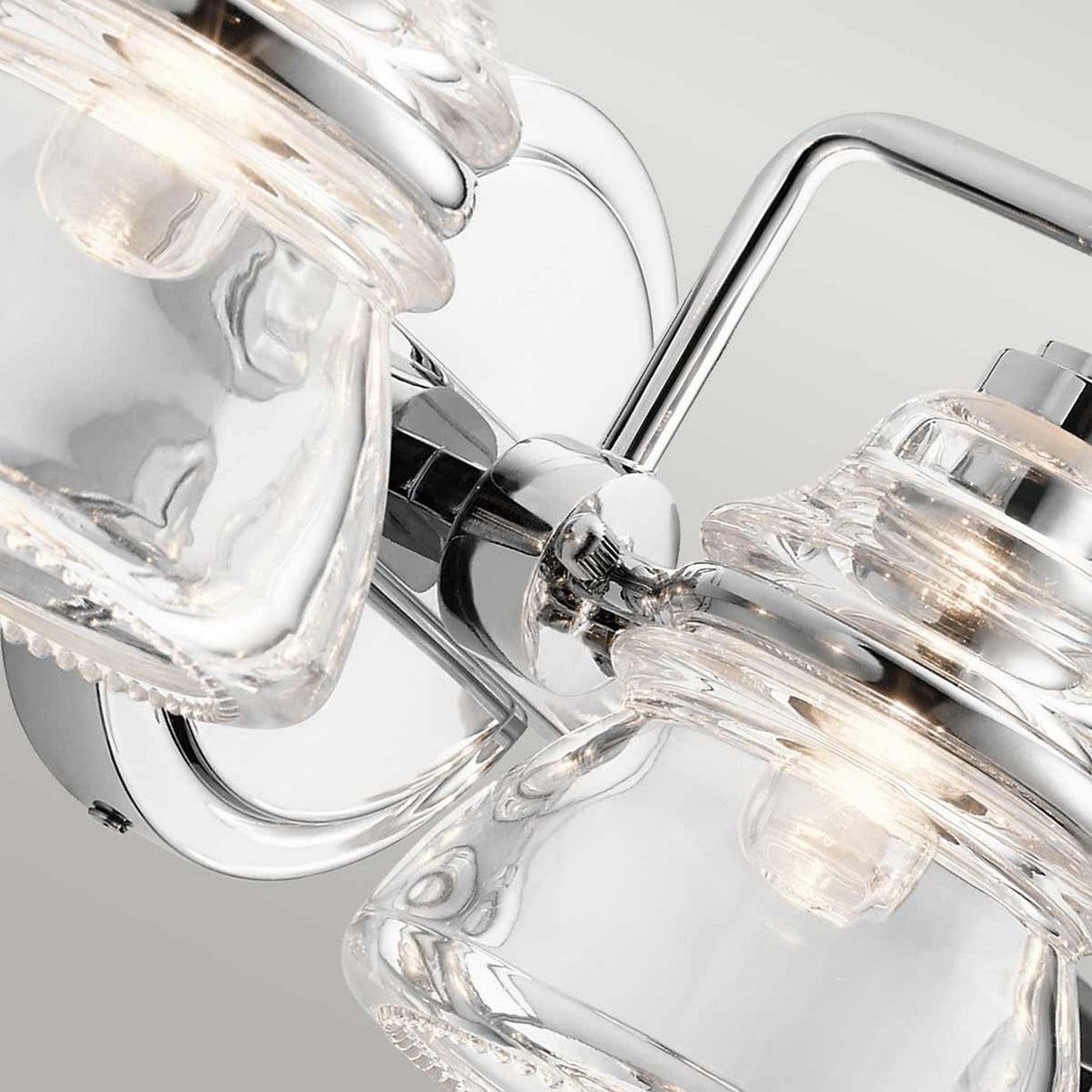 A close-up of the Talland 3 Light Wall Light - Polished Chrome reveals two visible bulbs within clear glass shades, gracefully reflecting light. This fixture is mounted on a wall with a gray background, ideal for enhancing bathroom illumination.