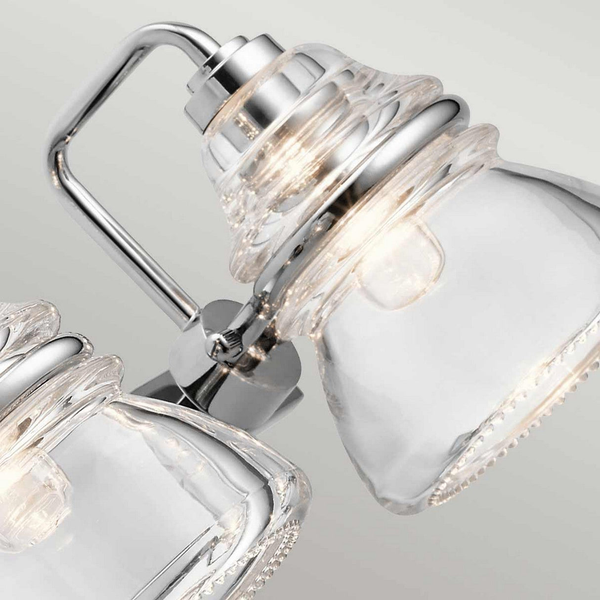 Close-up of two Talland 3 Light Wall Lights in polished chrome with clear bell-shaped shades. Elegantly designed, they showcase visible bulbs inside the glass and emit a soft glow perfect for bathroom illumination against a neutral background.