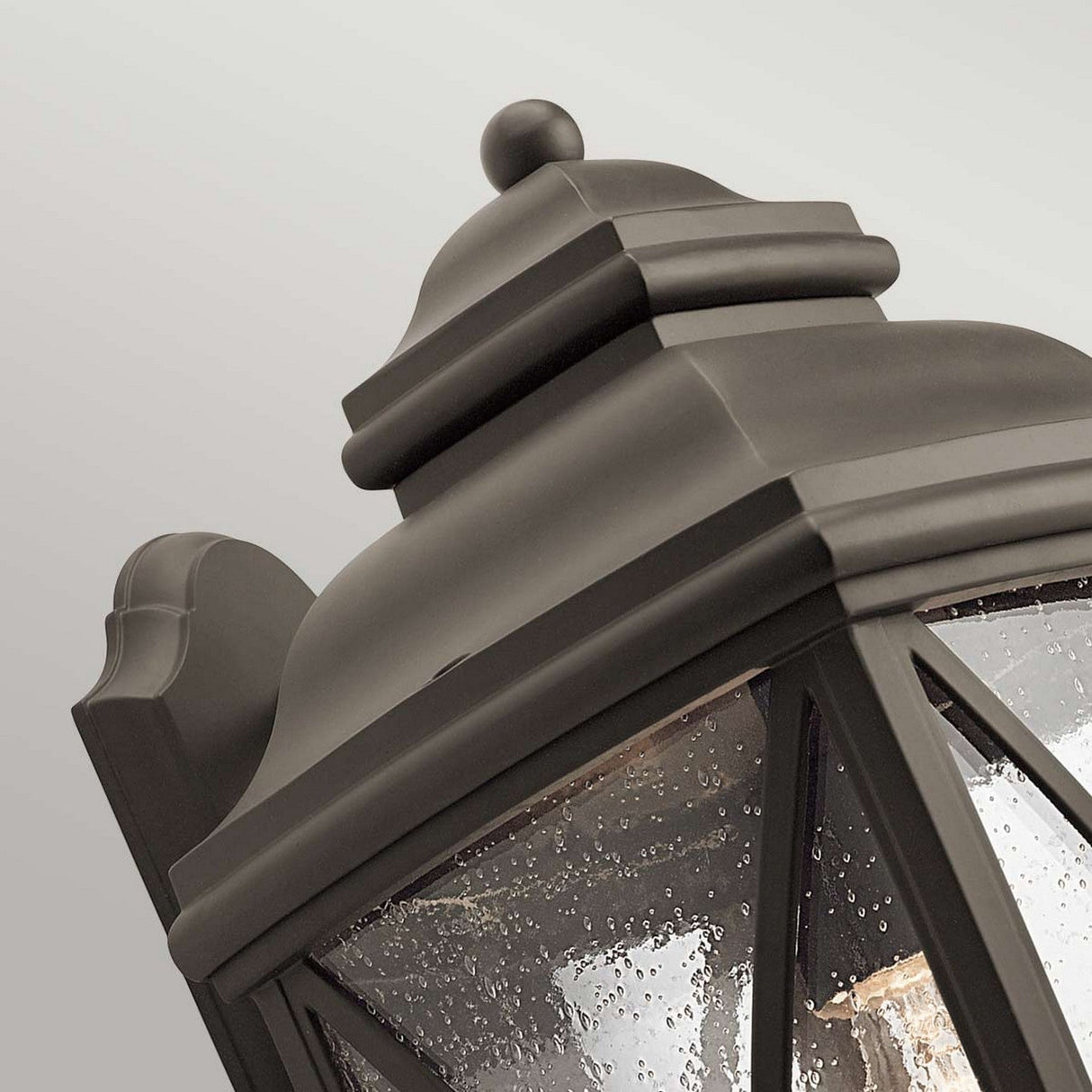 Close-up of a Tangier Outdoor 1 Light Large Wall Lantern in an Olde Bronze finish, featuring a classic design. The damp glass panels suggest rain, and the geometric patterns along with the ornamental top create a striking contrast against a gray background, exuding timeless elegance.