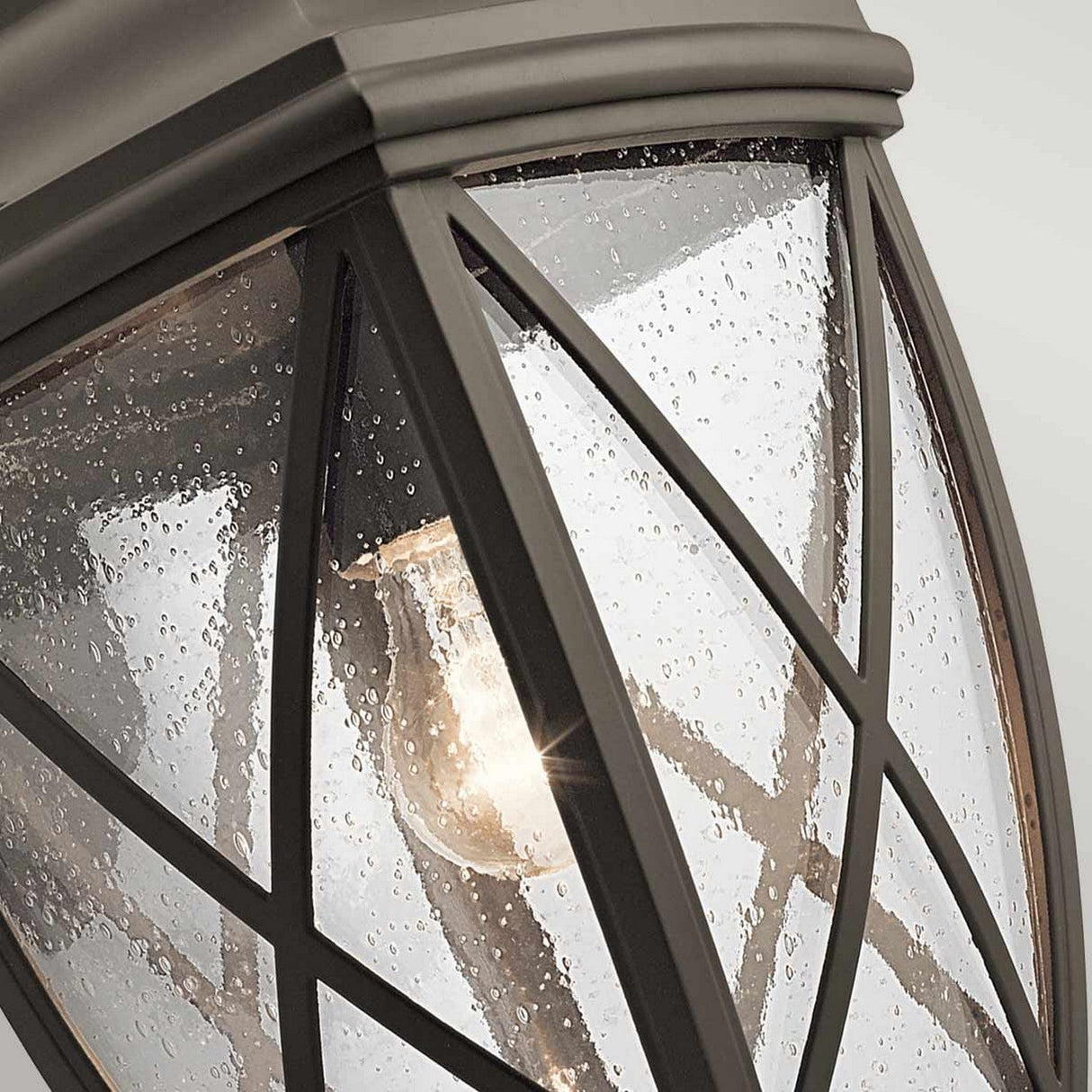 Introducing the Tangier Outdoor 1 Light Large Wall Lantern in Olde Bronze: This elegant piece highlights an exposed bulb encased within textured raindrop glass panels, all framed by a metal structure exhibiting an intersecting pattern. The glowing bulb emits a warm ambiance that evokes the essence of a Tangier Lantern.