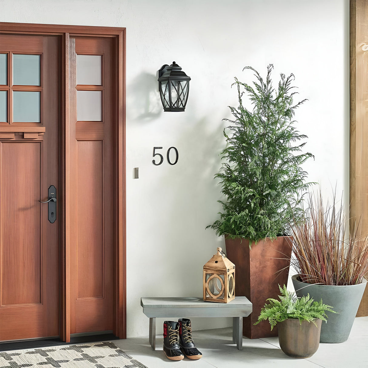 You'll find a cozy entryway featuring a wooden front door with glass panels, complemented by the Tangier Outdoor 1 Light Medium Wall Lantern in an Olde Bronze finish. A small bench holds boots and a decorative lantern, while potted plants and a rug complete the welcoming scene, all enhanced by the wall lantern's IP44 rating for durability.