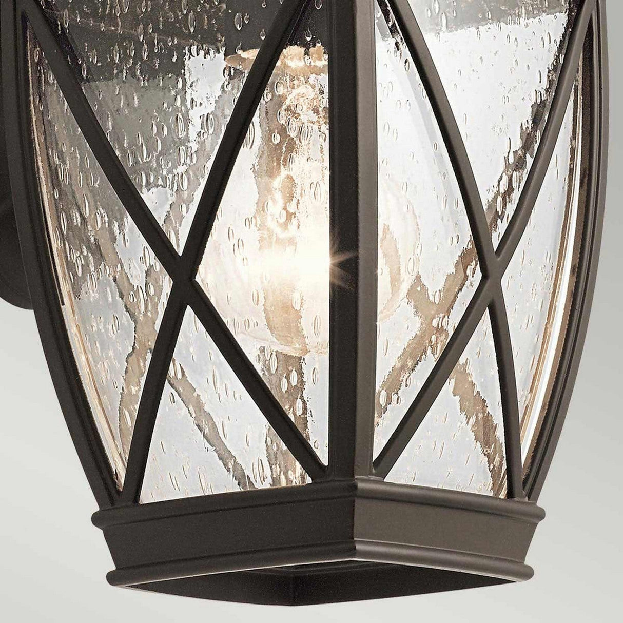 Close-up of the Tangier Outdoor 1 Light Small Wall Lantern in Olde Bronze, featuring a black metal frame and clear glass panels with a textured bubbly effect. The glowing bulb inside casts a warm light, complementing its elegant and modern design. This lantern is also IP44 rated for weather resistance.
