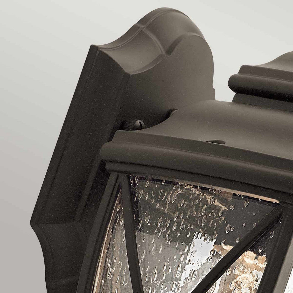 Close-up of the Tangier Outdoor 1 Light Small Wall Lantern, showcasing its Olde Bronze black metal frame and glass panel. This outdoor lighting piece features droplets on textured glass, evoking a rainy atmosphere, while the decorative curves and geometric patterns of the frame add to its appeal.
