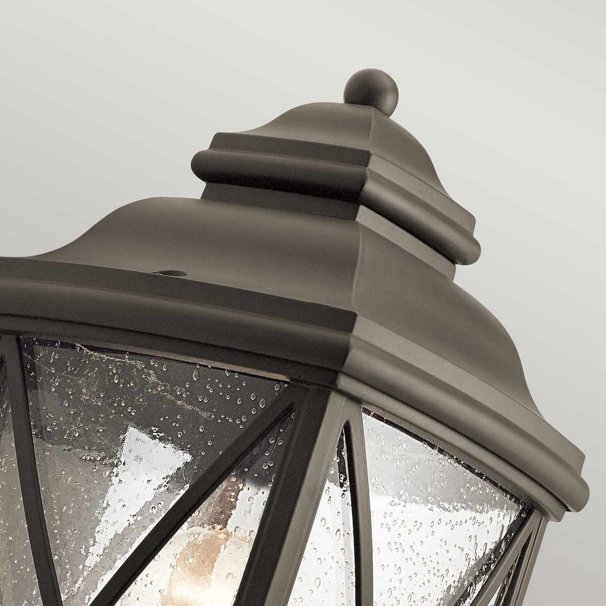 A close-up of the Tangier Outdoor 1 Light Medium Pedestal in Olde Bronze reveals its black metal with a peaked top and rain-speckled glass panels. The geometric crisscross frame is visible, beautifully contrasting against the plain light gray background.