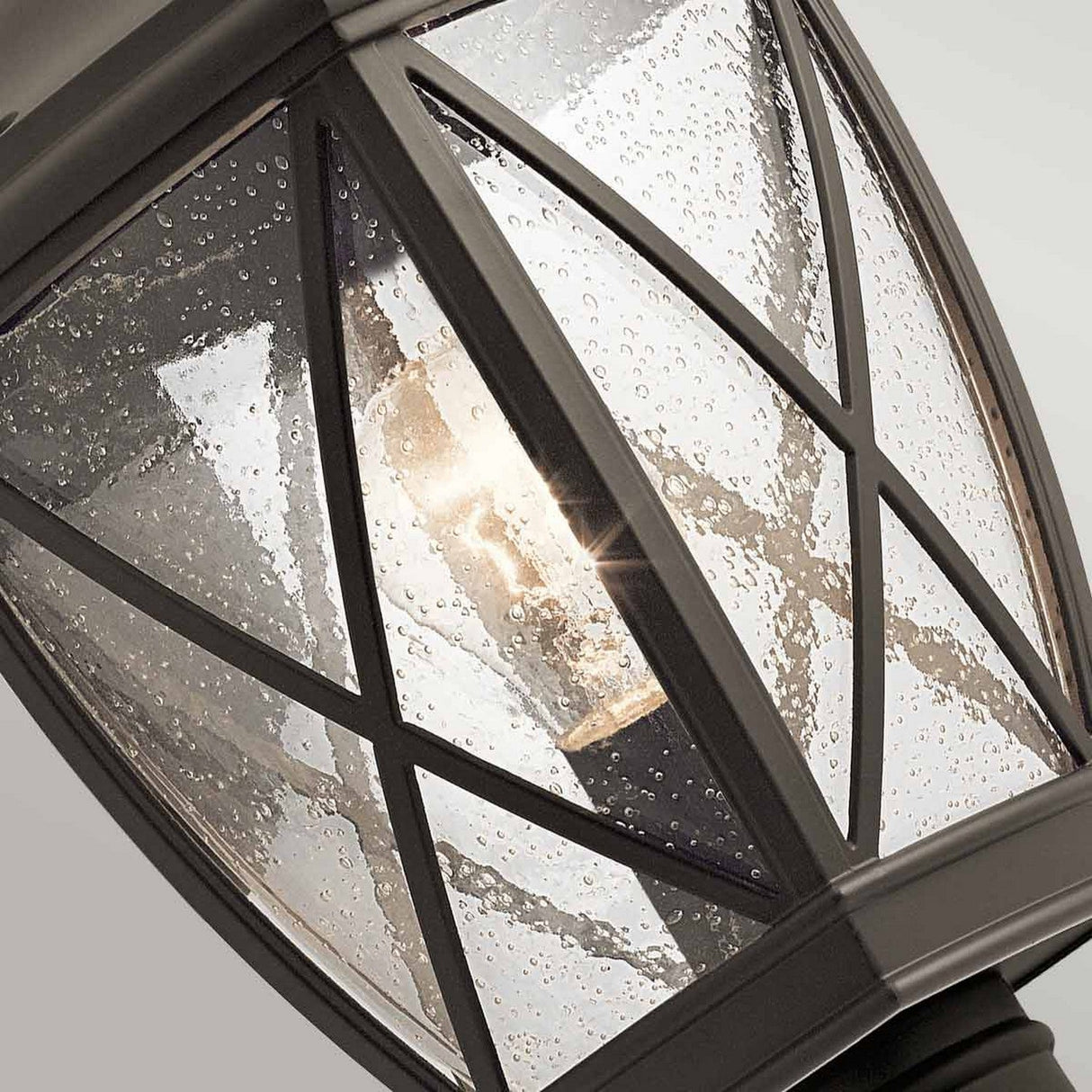 Close-up of the Tangier Outdoor 1 Light Medium Pedestal - Olde Bronze, featuring raindrop-patterned glass panels. The crisscross frame with an Olde Bronze finish encases a glowing bulb, casting a warm glow against the textured glass.