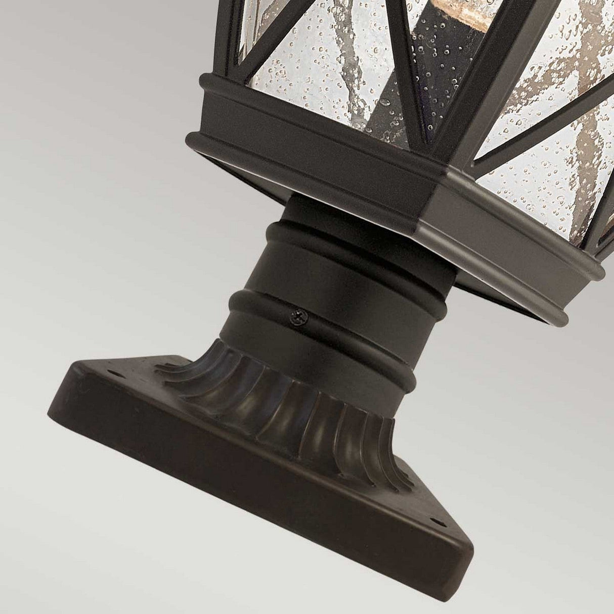 Close-up of the Tangier Outdoor 1 Light Medium Pedestal in Olde Bronze, featuring a black lantern with a textured glass design. This fixture includes a square base for mounting and a crisscross metal frame around the glass, offering a classic and decorative appearance.