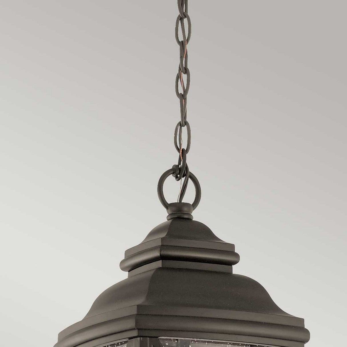 A close-up of the Tangier Outdoor 1 Light Medium Chain Pendant reveals its dark metal frame with an Olde Bronze finish and crisscross design. The pendant, with its decorative clear glass panels, hangs elegantly against a plain gray background.