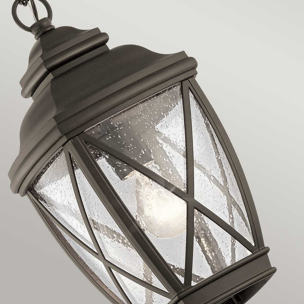 A close-up of the Tangier Outdoor 1 Light Medium Chain Pendant in Olde Bronze highlights its classic black metal design with a crisscross frame. The lantern showcases clear glass panels that reveal a visible light bulb inside, and the Olde Bronze finish accentuates droplets of water on the glass, suggesting recent rain.
