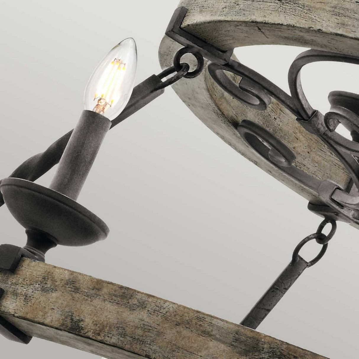 A close-up of the Taulbee 15 Light Chandelier - Weathered Zinc highlights its circular wooden frame with weathered zinc accents. A single exposed bulb emits a soft glow, suspended by a metal arm, artfully combining industrial and vintage elements to epitomize rustic aesthetics.