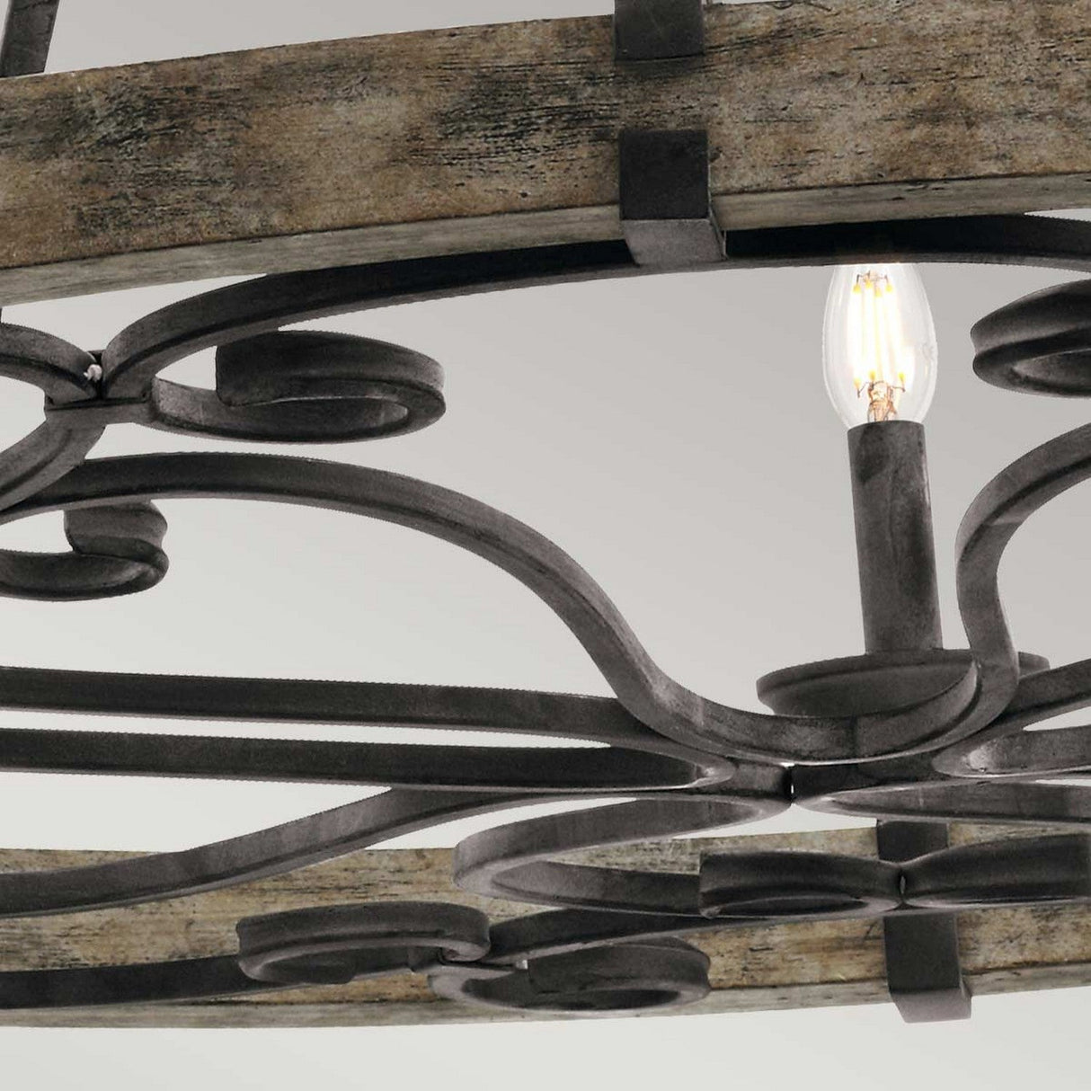 Close-up of the Taulbee 15 Light Chandelier, showcasing its intricate wrought iron design with a single illuminated candle-style bulb. The weathered zinc metalwork contrasts beautifully with the distressed wooden elements, enhancing its vintage industrial aesthetic.