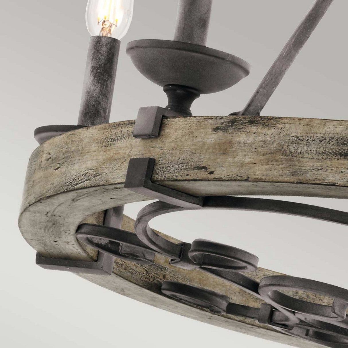 A close-up of the Taulbee 6 Light Chandelier - Weathered Zinc reveals its circular wooden frame adorned with weathered zinc accents. One illuminated candle-shaped bulb stands out against a light gray background, perfectly blending industrial and vintage elements.