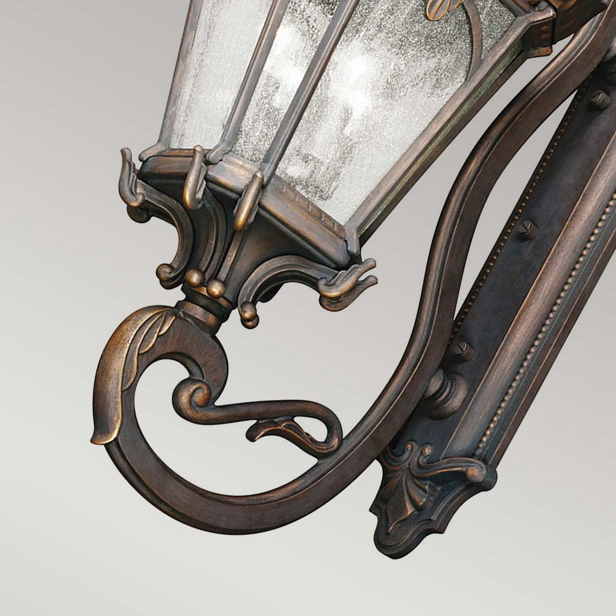 A detailed view of the Tournai Outdoor 4 Light Grand Extra Large Wall Lantern showcases its intricate, vintage-inspired design, with a gracefully curved, decorative metal framework and seedy glass enclosure. The metal boasts an antique bronze finish that gleams against a simple, light gray background.