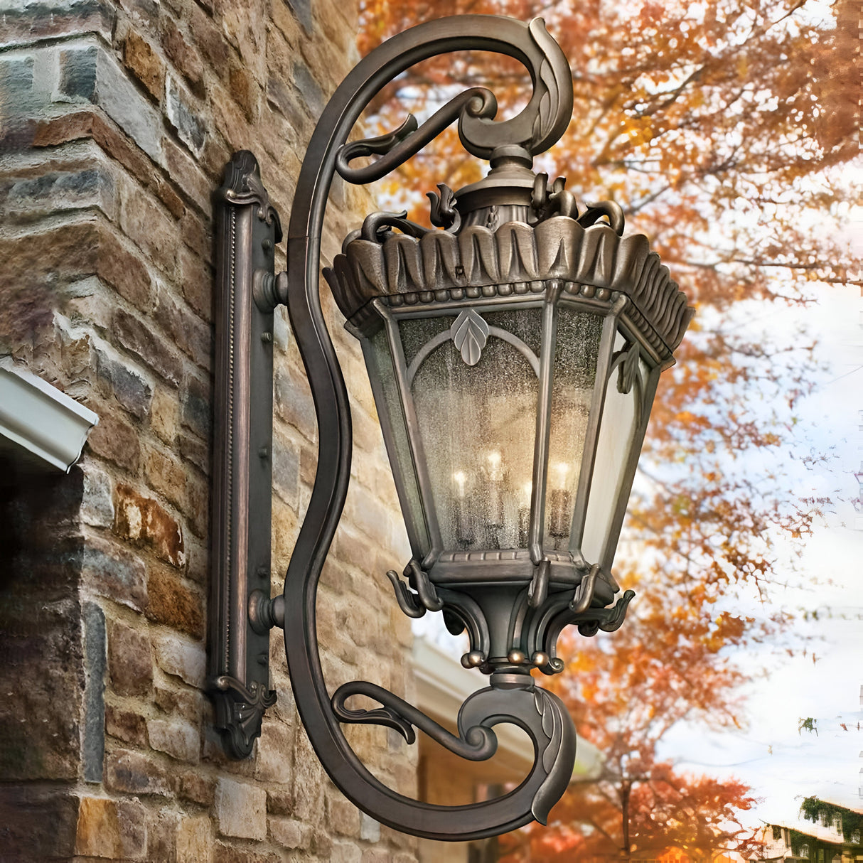 The Tournai Outdoor 4 Light Grand Extra Large Wall Lantern in a bronze finish is mounted on a stone wall, showcasing its intricate metal design that frames seedy glass panels softly glowing with light. In the background, autumn leaves add a warm, seasonal atmosphere.