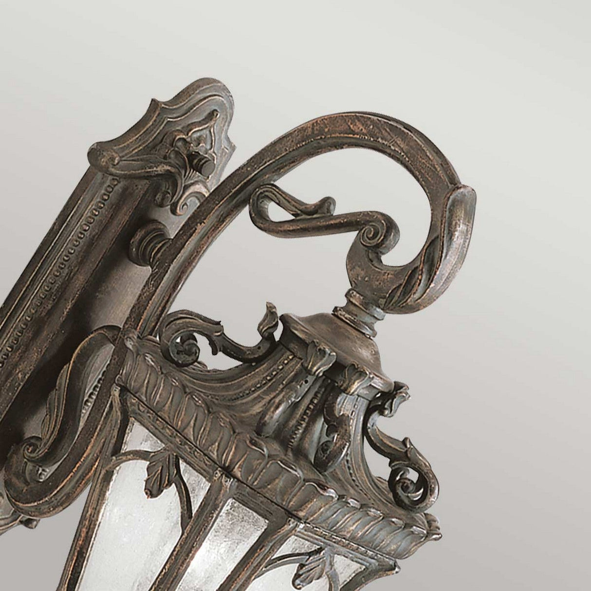 A close-up of the Tournai Outdoor 2 Light Large Wall Lantern in bronze showcases its ornate vintage-style design, with intricate metalwork and glass panels. Mounted on a textured wall, this lamp features a decorative finial and curved bracket, evoking a historic lantern design and classic, elegant aesthetic.