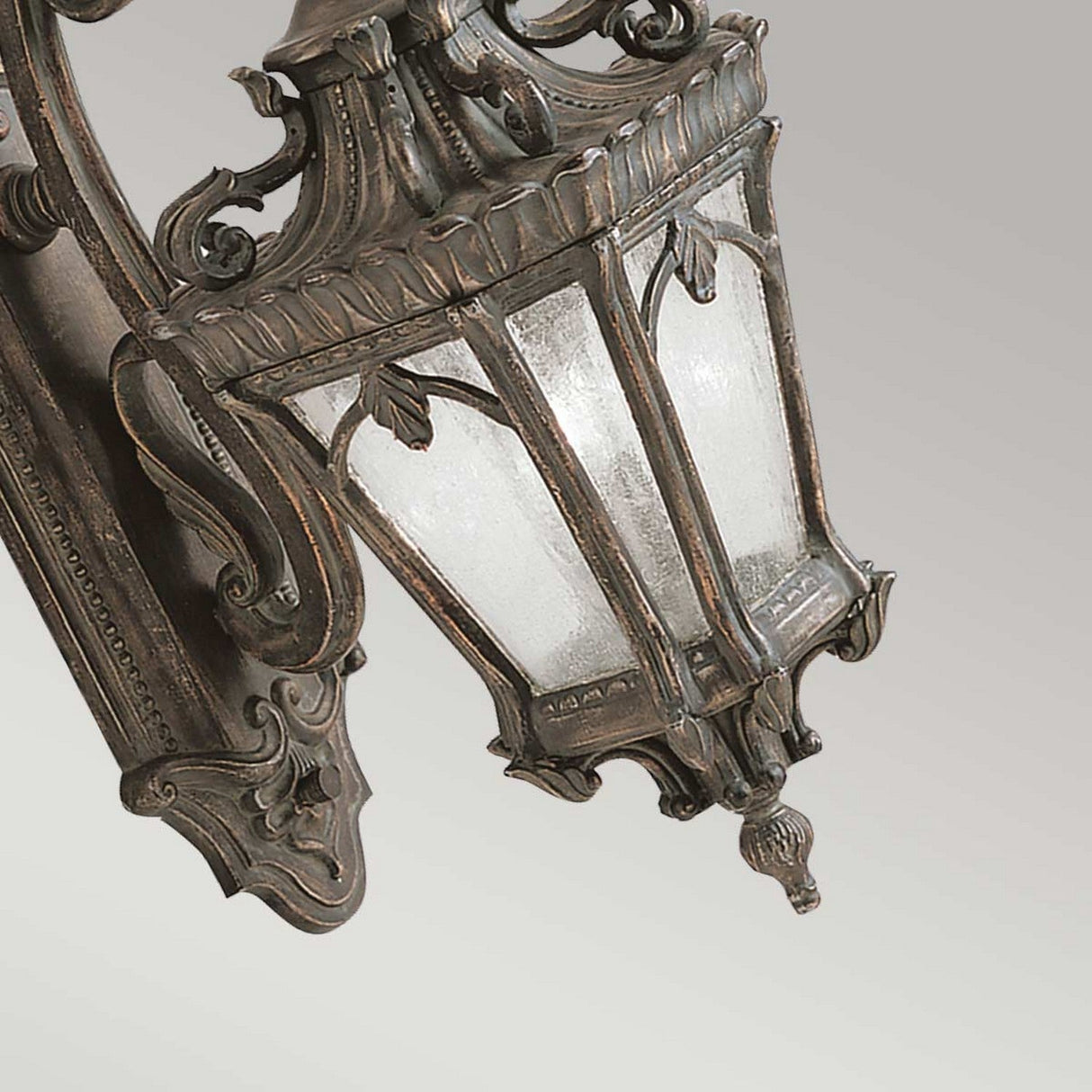 The Tournai Outdoor 2 Light Large Wall Lantern in Bronze, featuring intricate metalwork and frosted glass panels, hangs against a gradient gray background. This lantern exudes an antique touch with its ornate details and dark bronze finish.