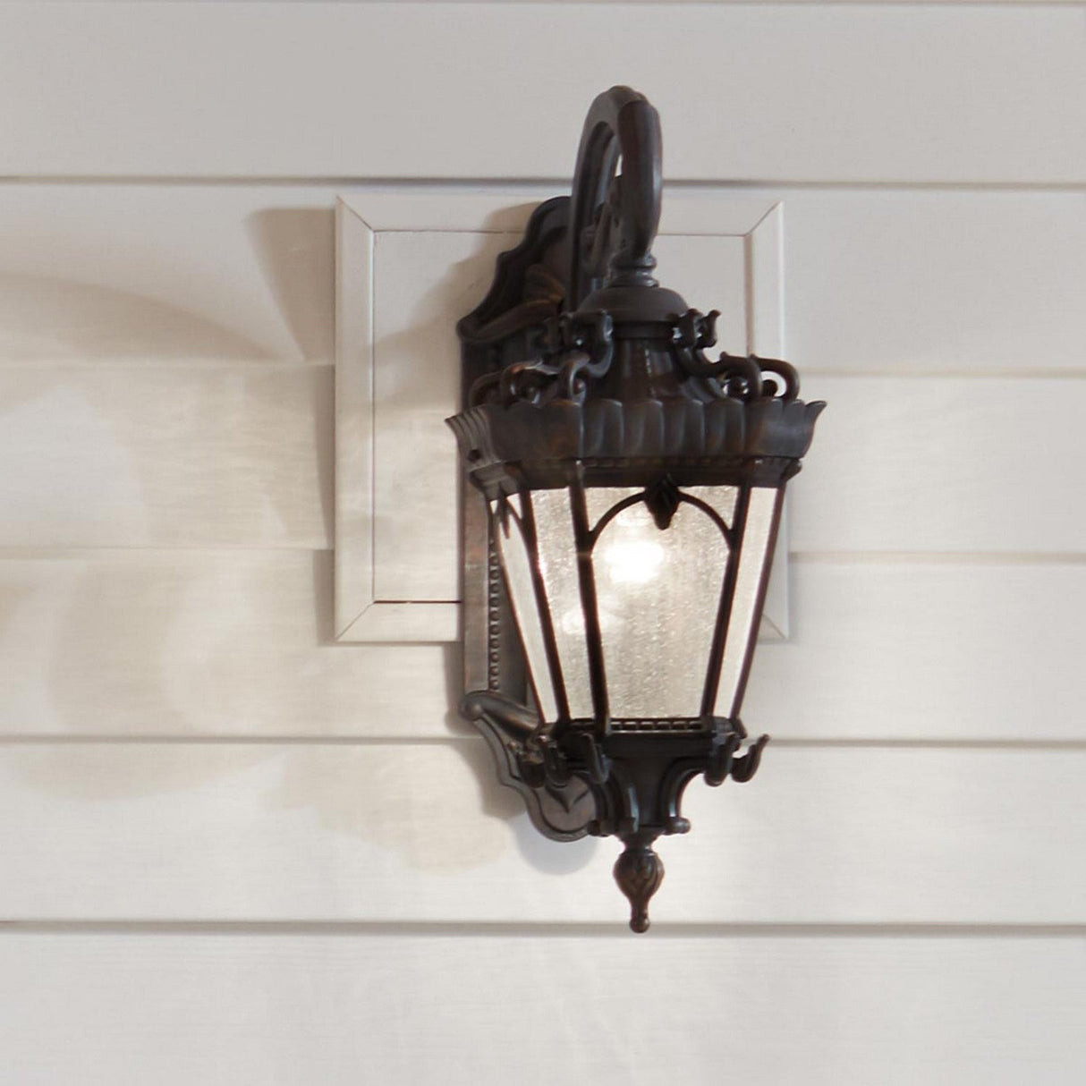 A Tournai Outdoor 2 Light Large Wall Lantern in bronze, with a vintage-style black metal design and glass enclosure, is mounted on a white-paneled exterior wall. The lantern showcases intricate historic designs and emits a warm glow when lit.